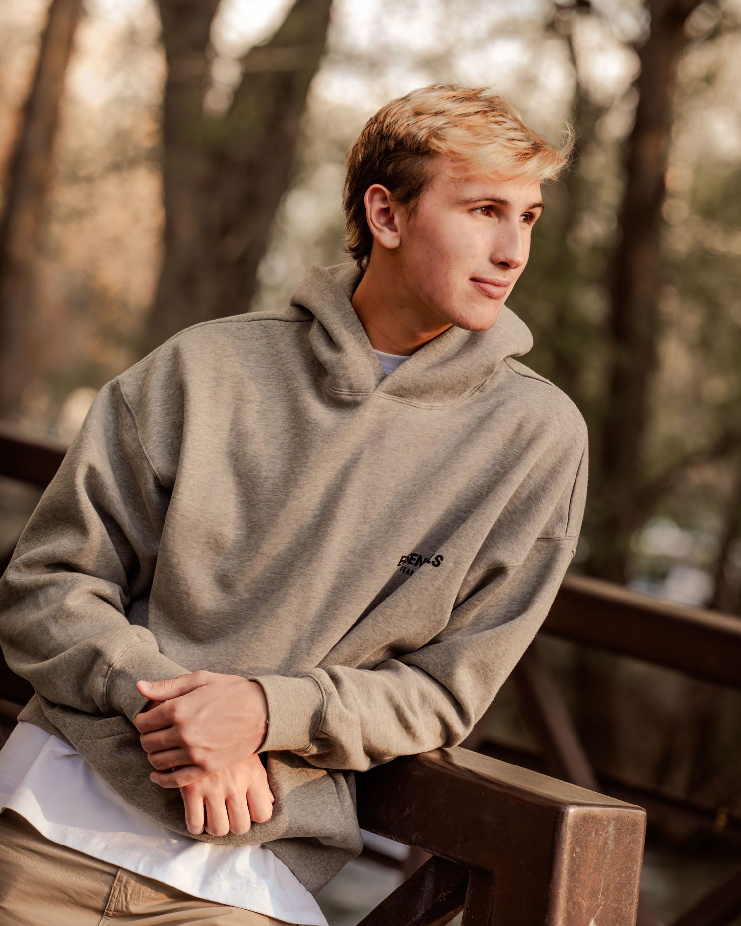 Young man in gray hoodie sitting outdoors with blurred trees in background, casual fashion portrait.