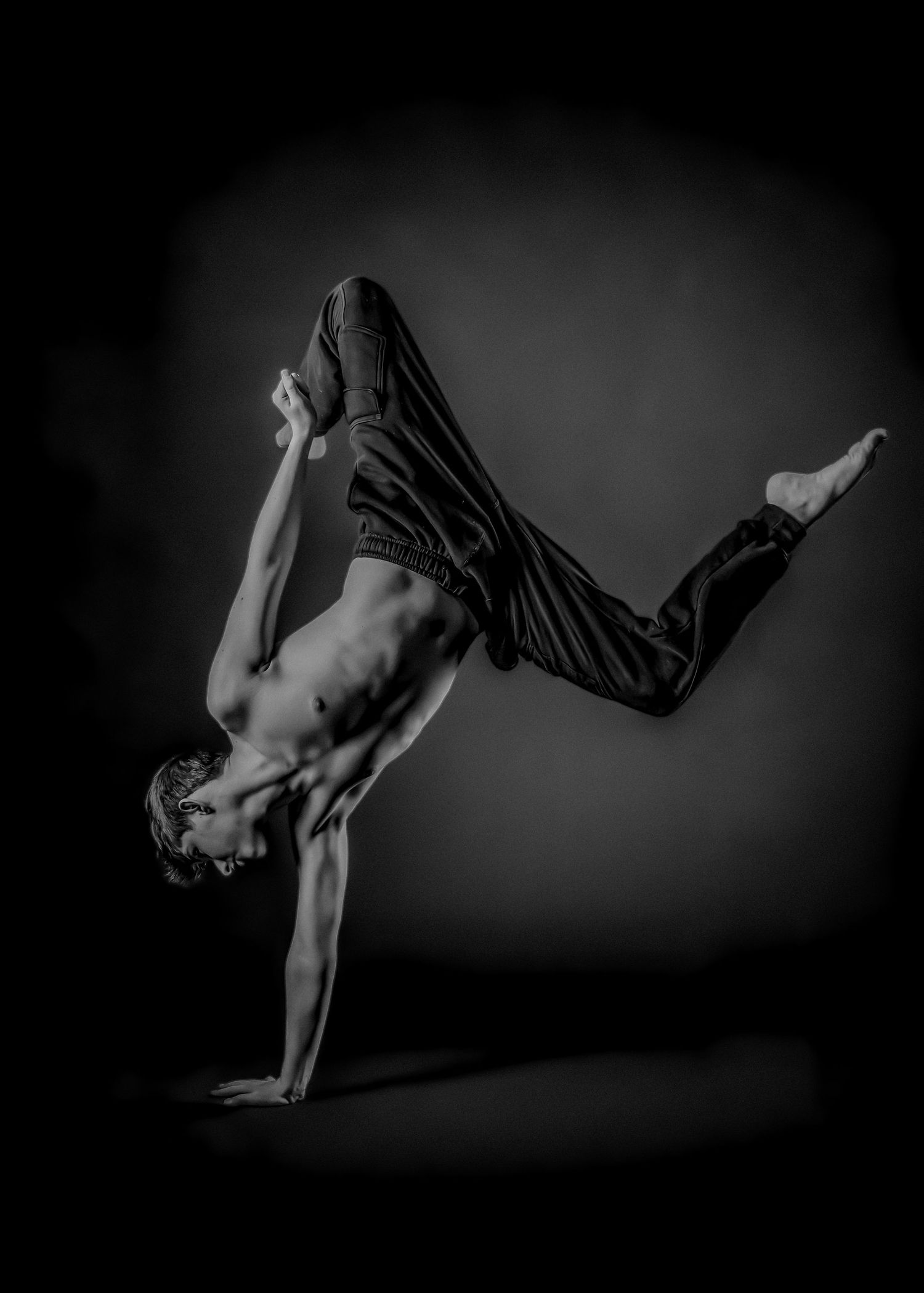 A dancer performs a dynamic one-arm handstand pose in black and white against a dark background.