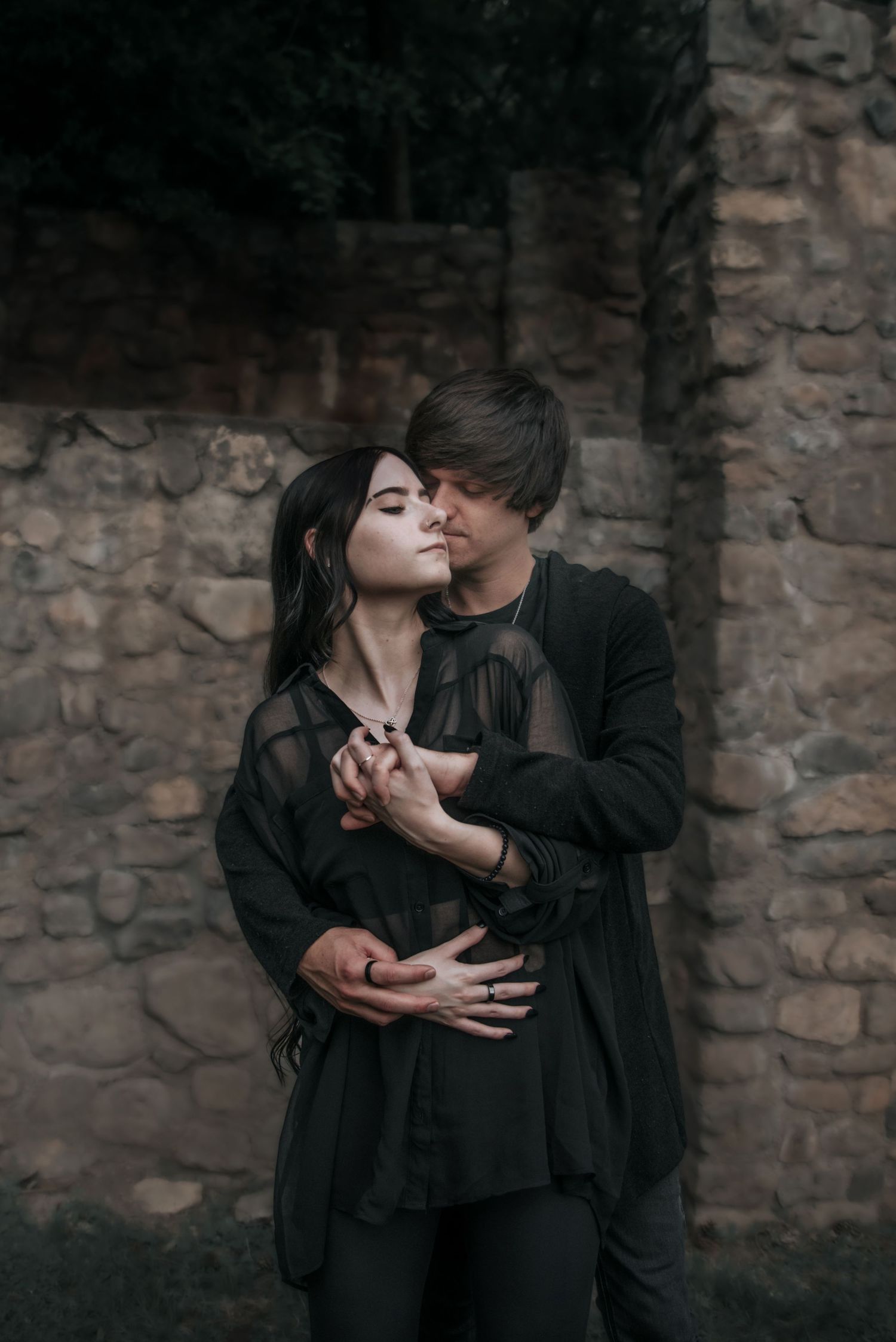 Romantic couple in all black sharing an intimate moment against a stone wall backdrop.