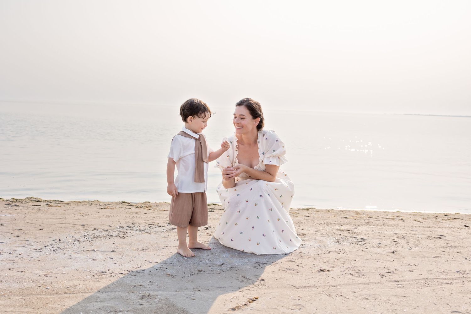 A mother and child in white outfits walks along a serene beach during sunset, creating silhouettes against the lake.