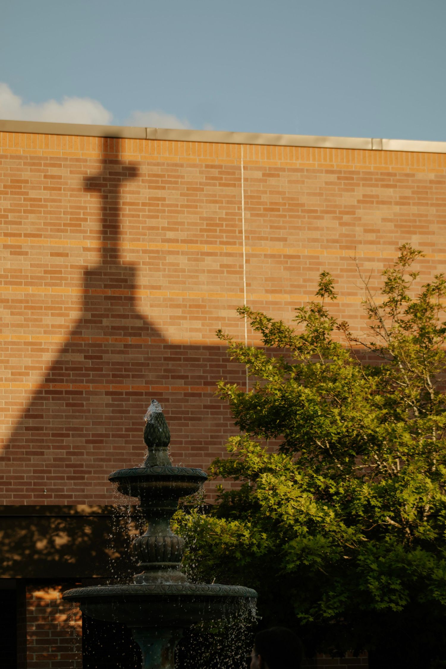 A cross shadow falls dramatically on a brick church exterior wall next to a garden fountain.