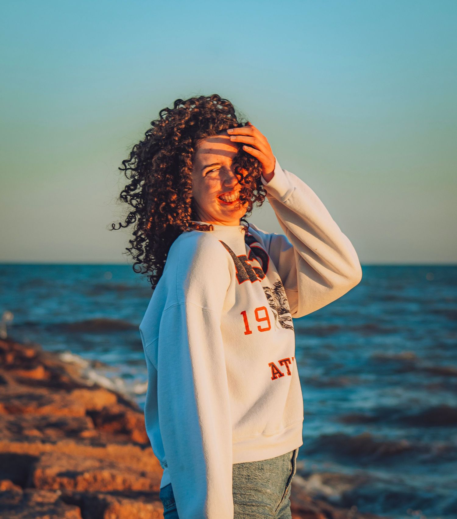 Young woman with curly hair in white athletic sweatshirt poses on rocky shoreline at golden hour sunset.
