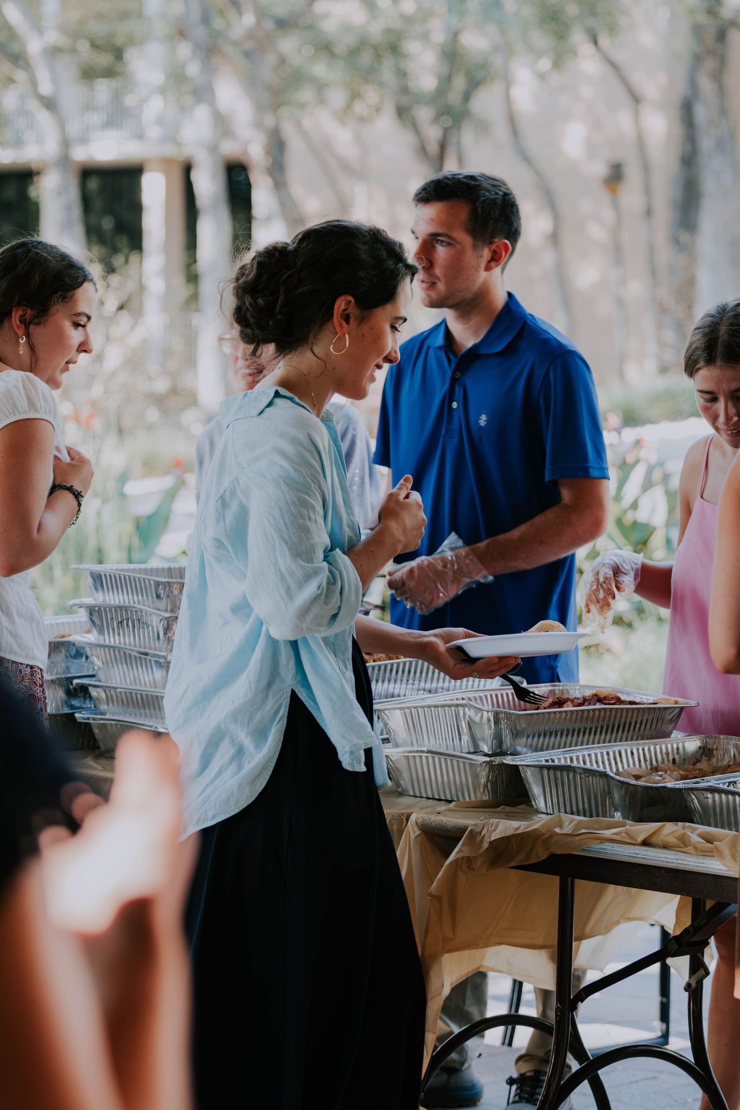 People serving food at an outdoor buffet table during a casual catered event in a garden setting.