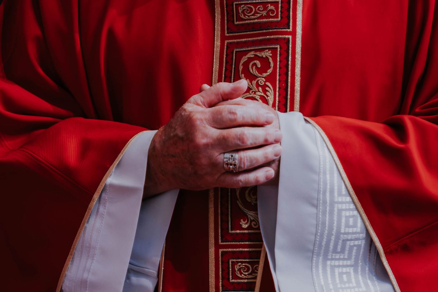 Close-up of a Catholic priest in red vestments with clasped hands wearing a ring during a religious ceremony.