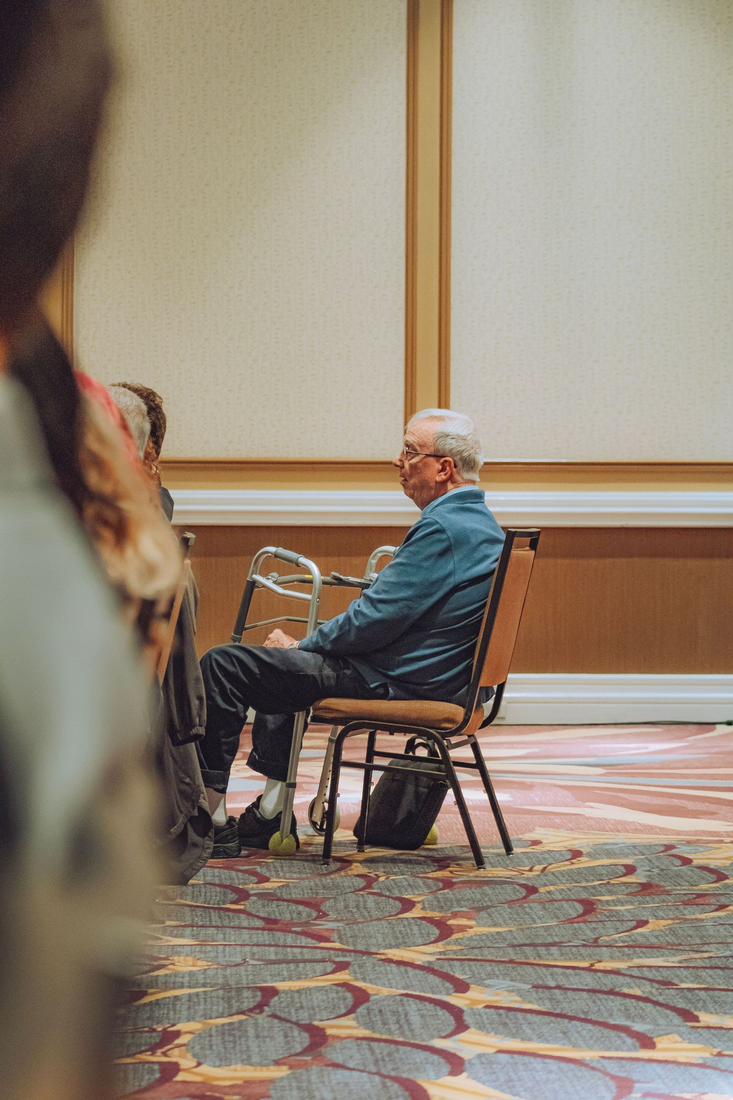 Elderly man sitting in chair at a formal indoor conference or seminar event.