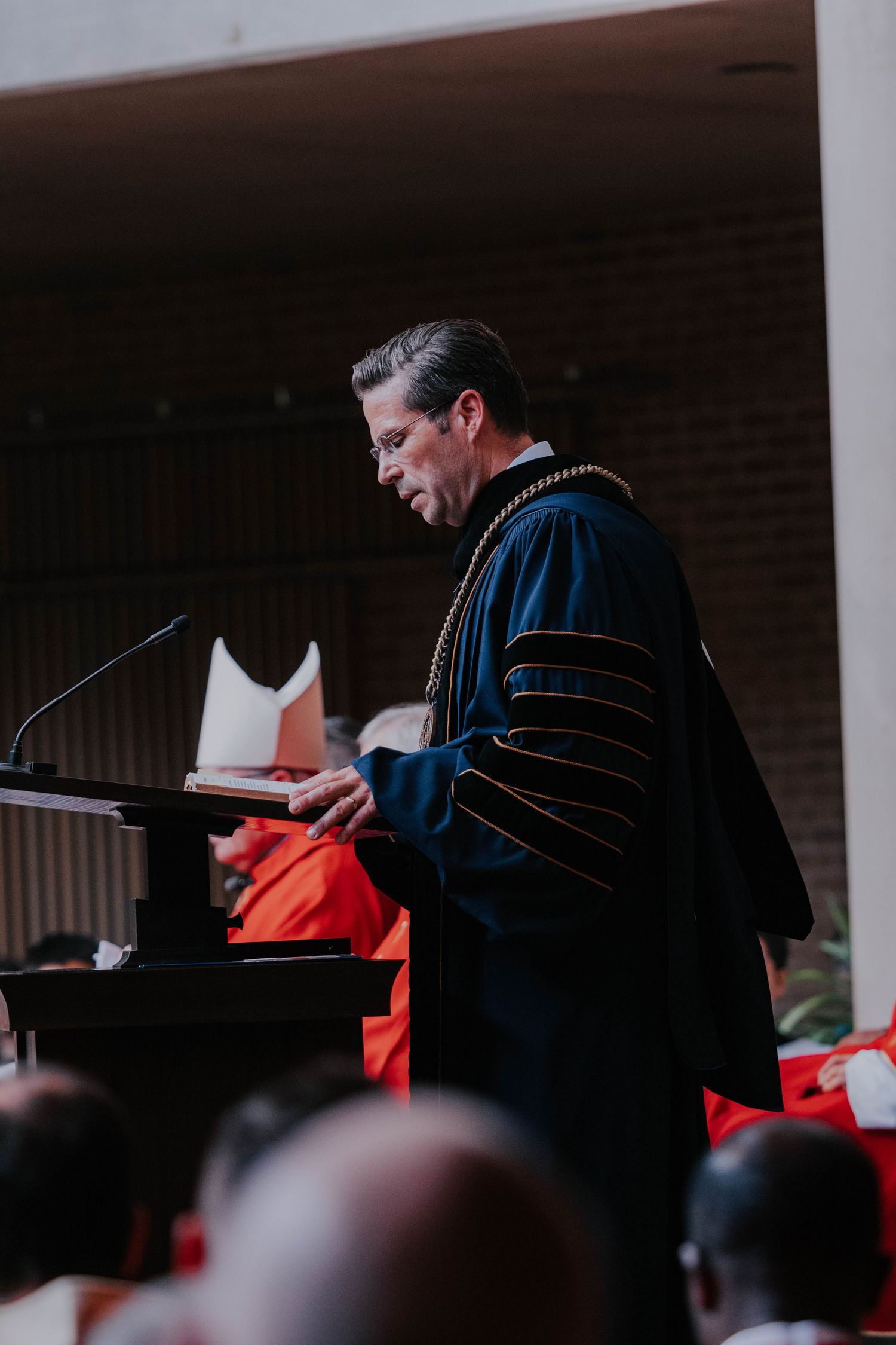 Academic official in doctoral regalia speaks at podium during graduation ceremony, with graduates in red robes seated behind.