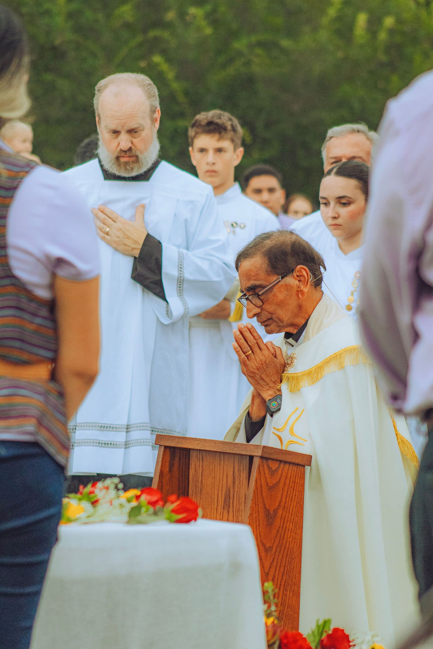 Catholic priest kneeling in prayer at an outdoor altar during a religious ceremony with attendees.