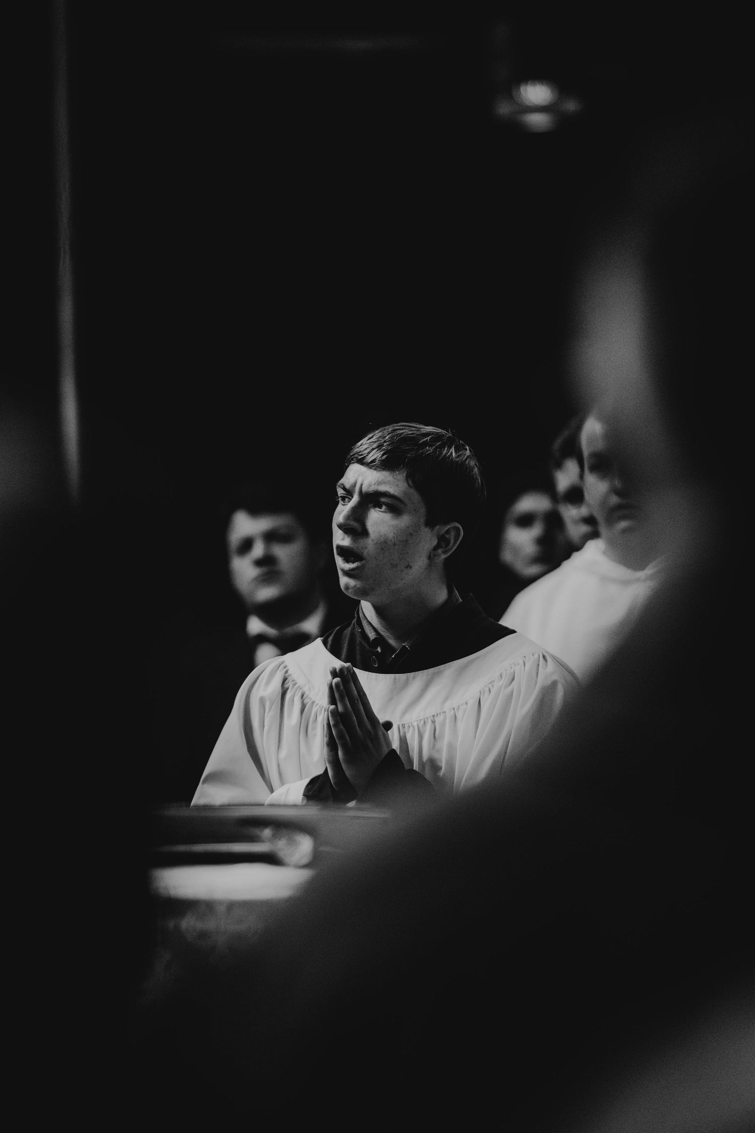 Young choir member in white robes praying with hands clasped in a dimly lit church setting.