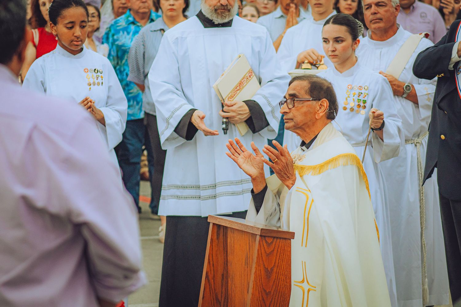 Catholic priest leads outdoor Mass at podium with altar servers and congregation gathered behind him.