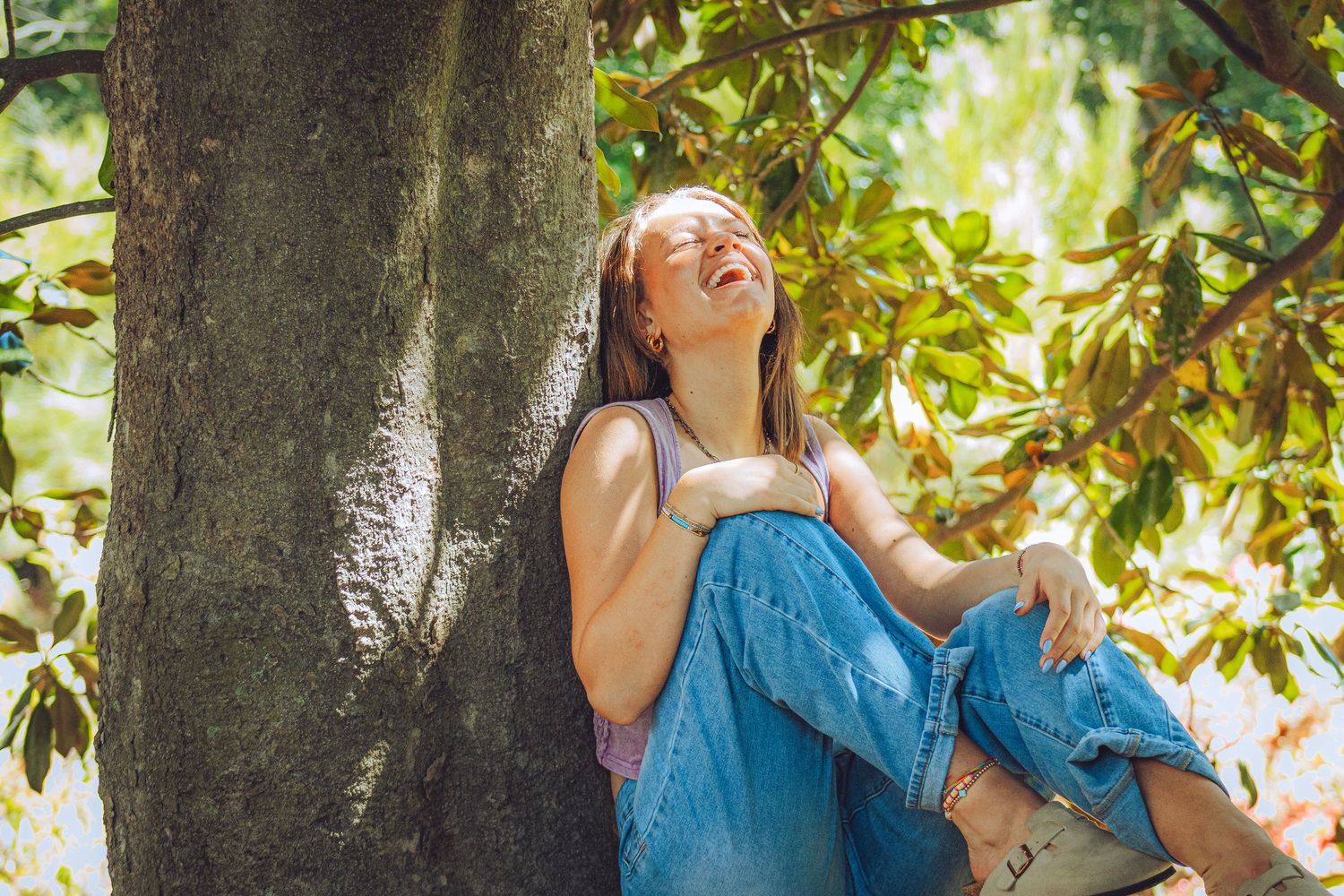 Happy young woman in jeans laughing while sitting against a tree trunk in lush green garden during her senior photo session.