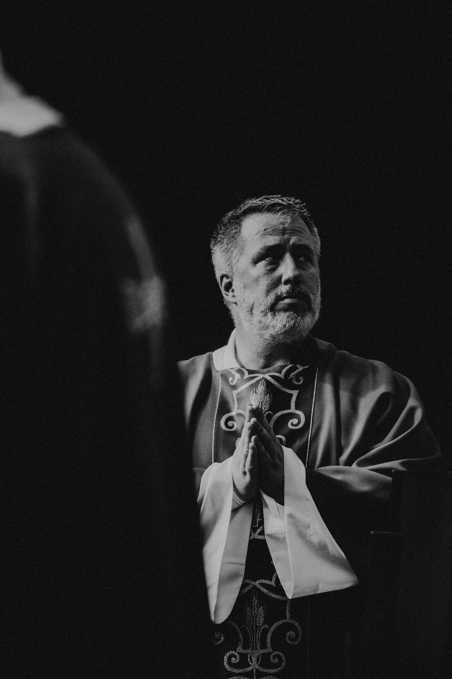 Black and white photo of a priest in vestments praying with hands clasped, looking upward in a dark church.