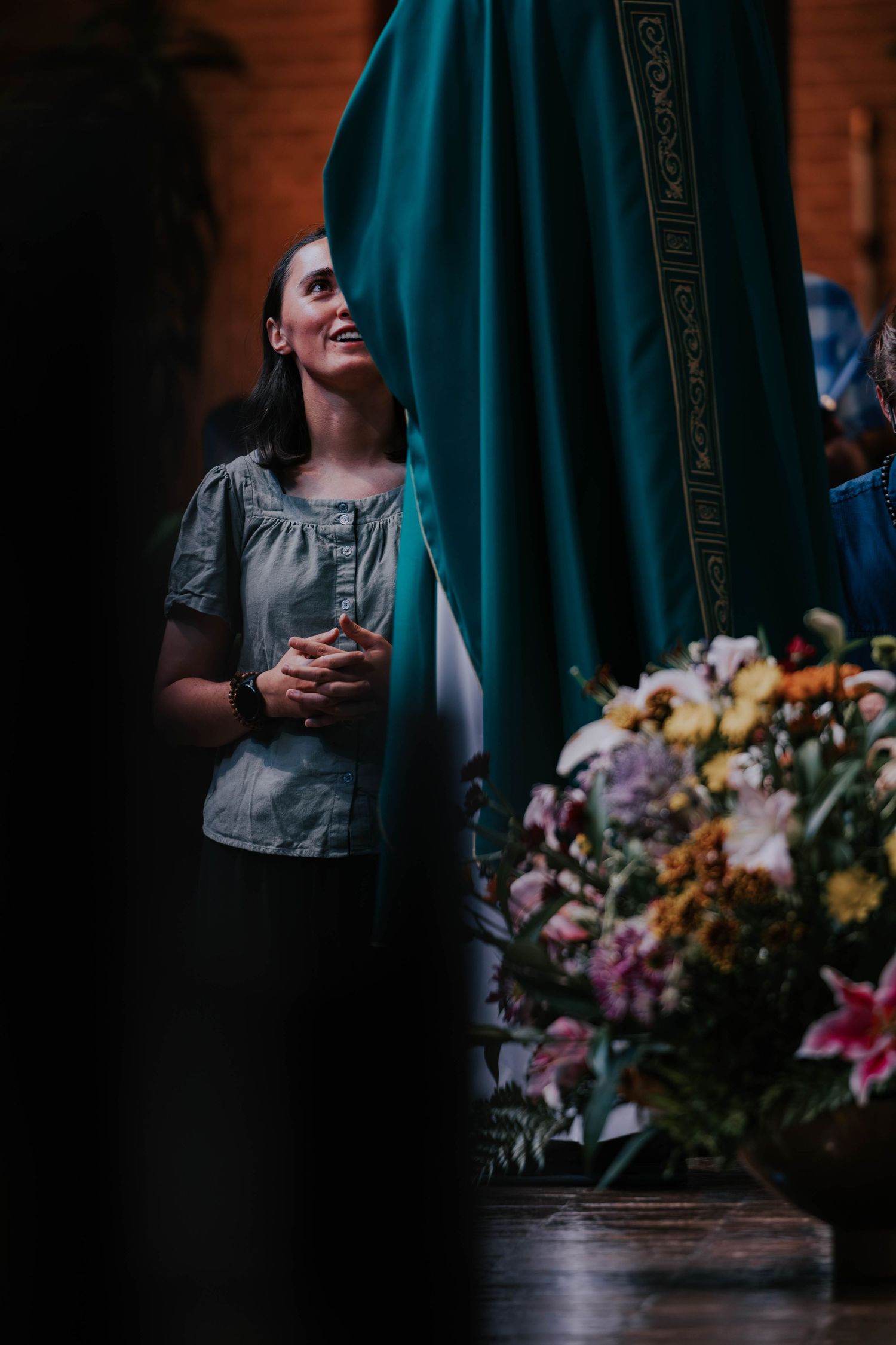 Woman in gray blouse praying near teal religious statue surrounded by colorful flowers in a church.