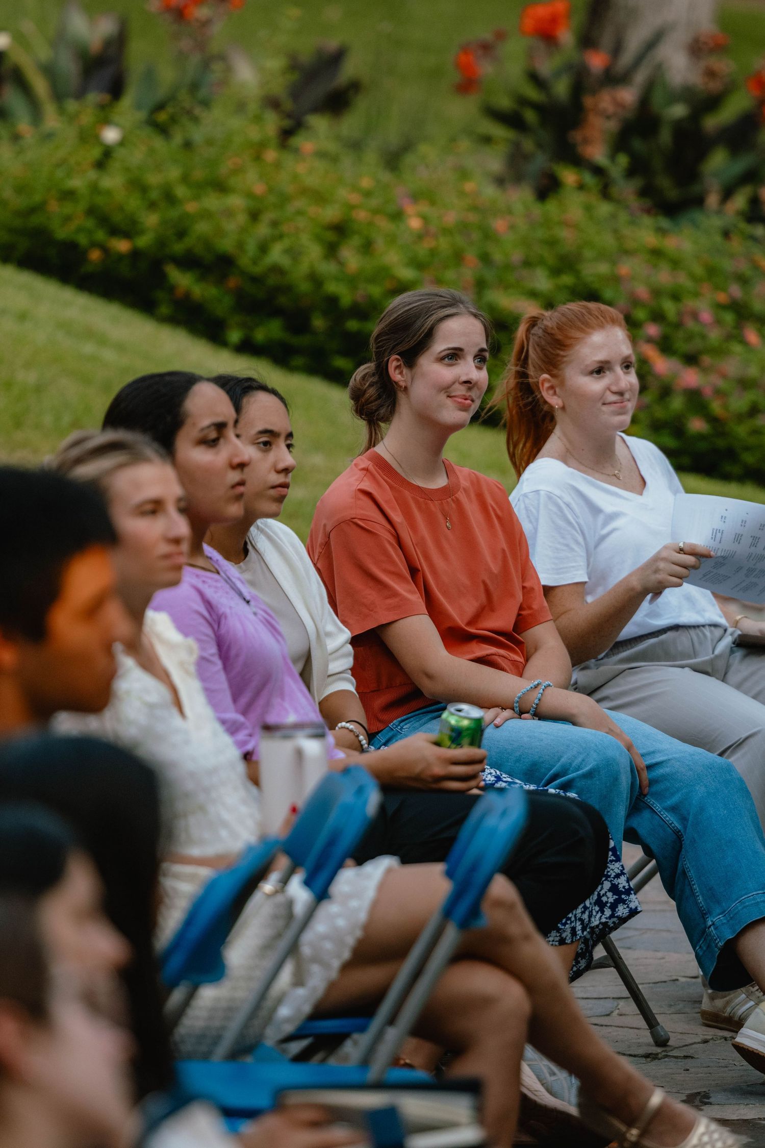 Group of young women sitting in chairs outdoors at a garden event, smiling and engaged in an outdoor gathering.