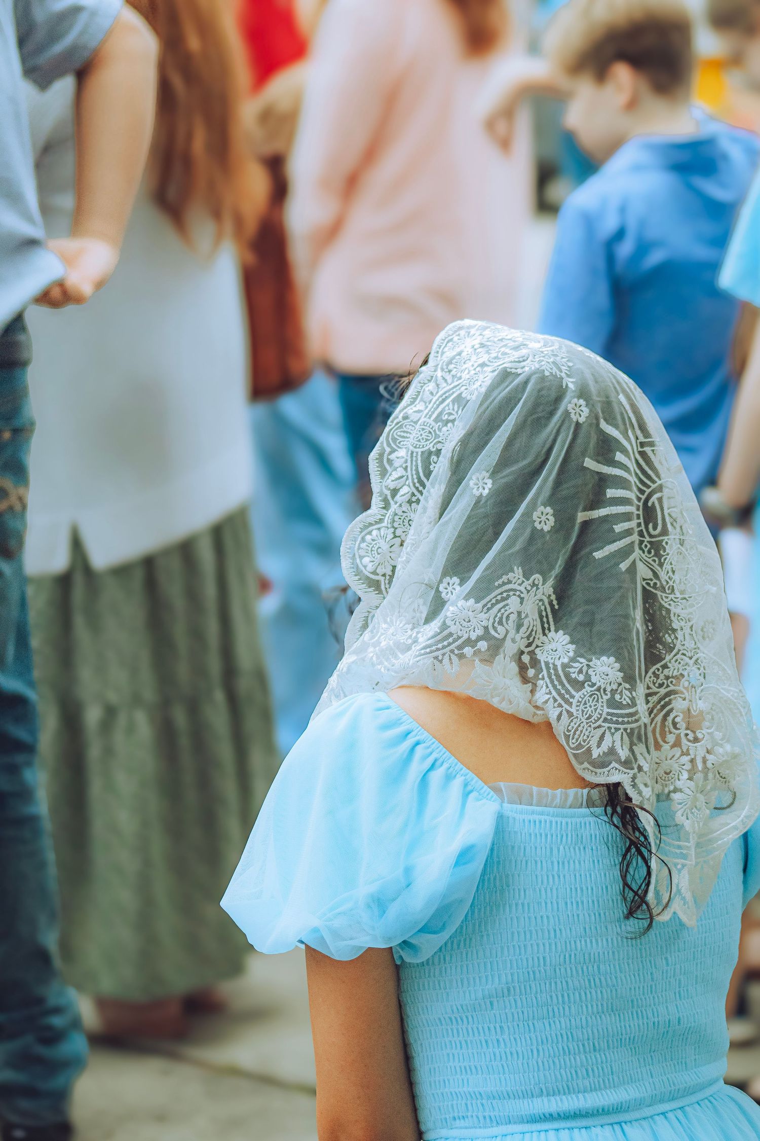 Young girl in blue dress wearing white lace chapel veil at an outdoor Catholic event.