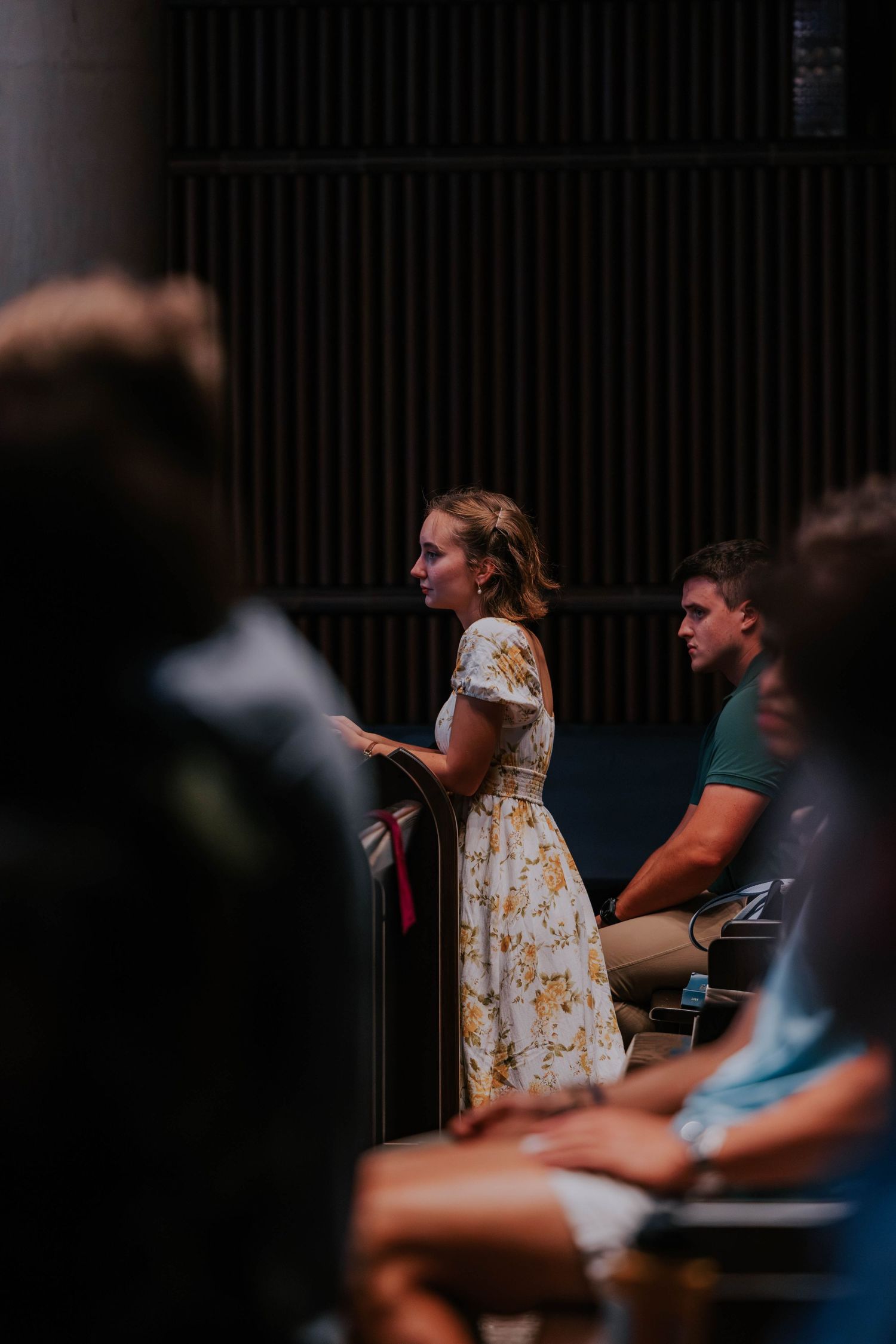 Young woman in floral dress standing in church pew during a service, with congregation members seated around her.