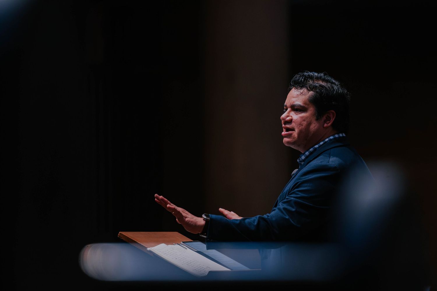 Man in dark suit speaking passionately at podium in dramatic low-light setting during formal presentation.