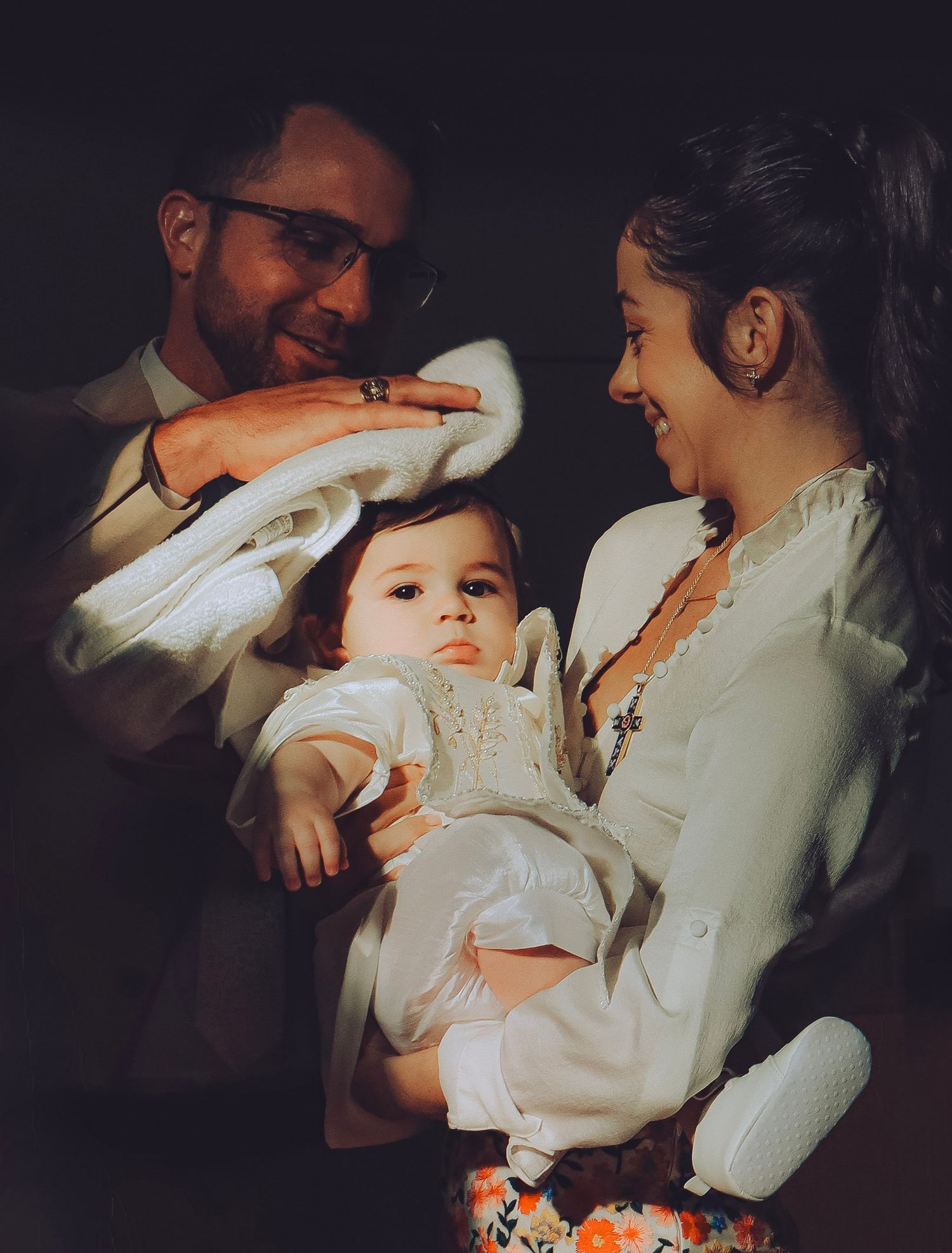 Smiling parents lovingly looking at their baby dressed in white during a baptism ceremony.