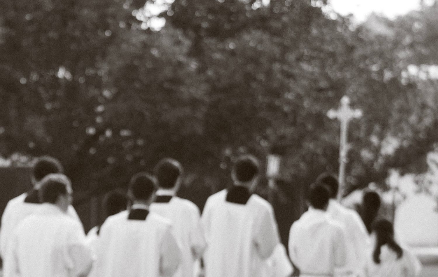 Black and white photo of robed clergy walking in procession toward a cross outdoors.