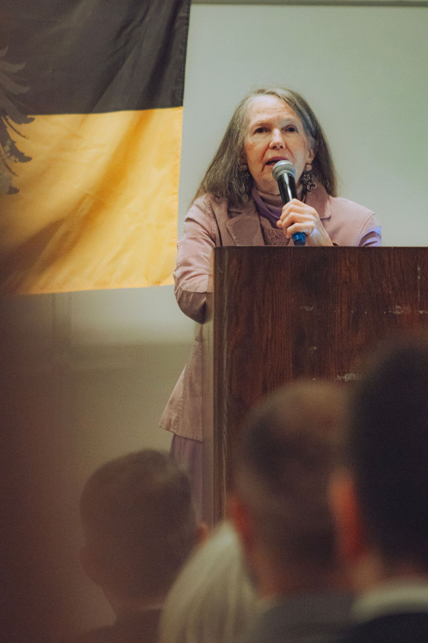 Elderly woman with long gray hair speaks at wooden podium with black and gold flag behind her at the Blessed Karl Symposium.