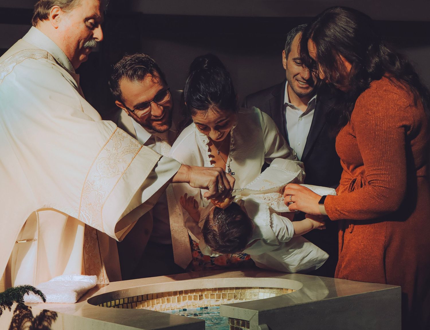 A priest performs a baptism, pouring water over an infant's head surrounded by family at the font.