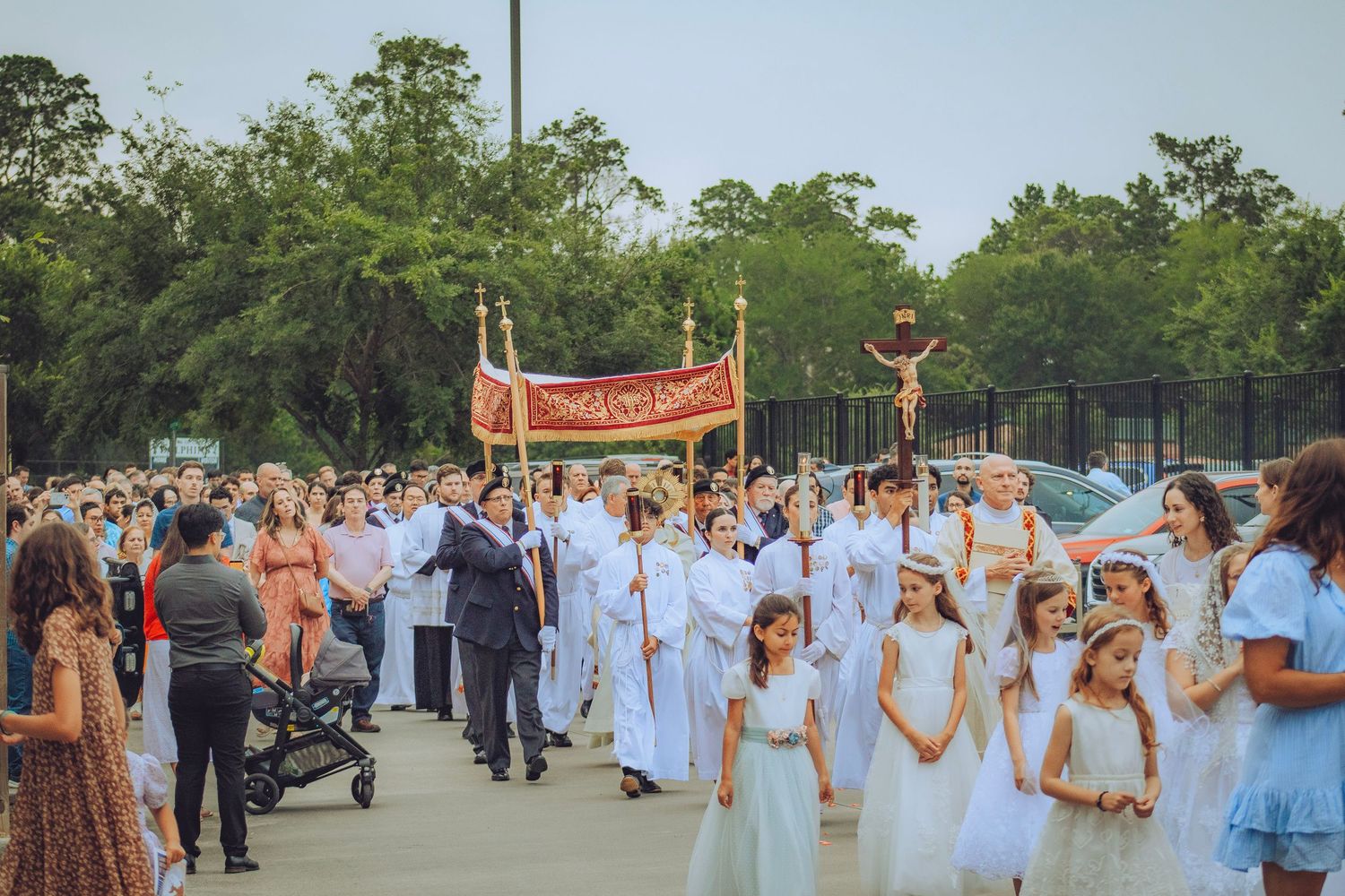 Catholic Corpus Christi procession with children in white and clergy carrying a red banner outdoors.