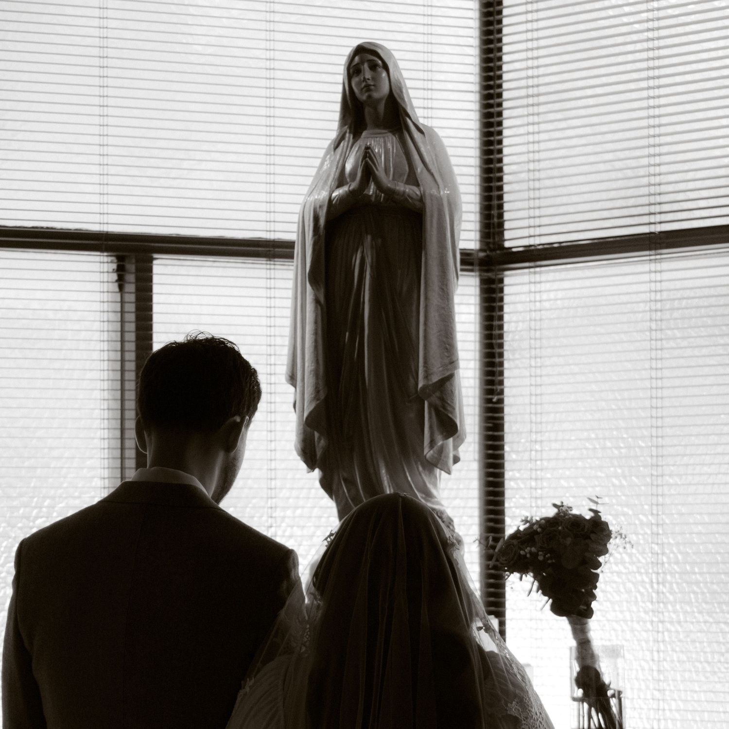 Bride and groom stand before Virgin Mary statue in black and white wedding photo.