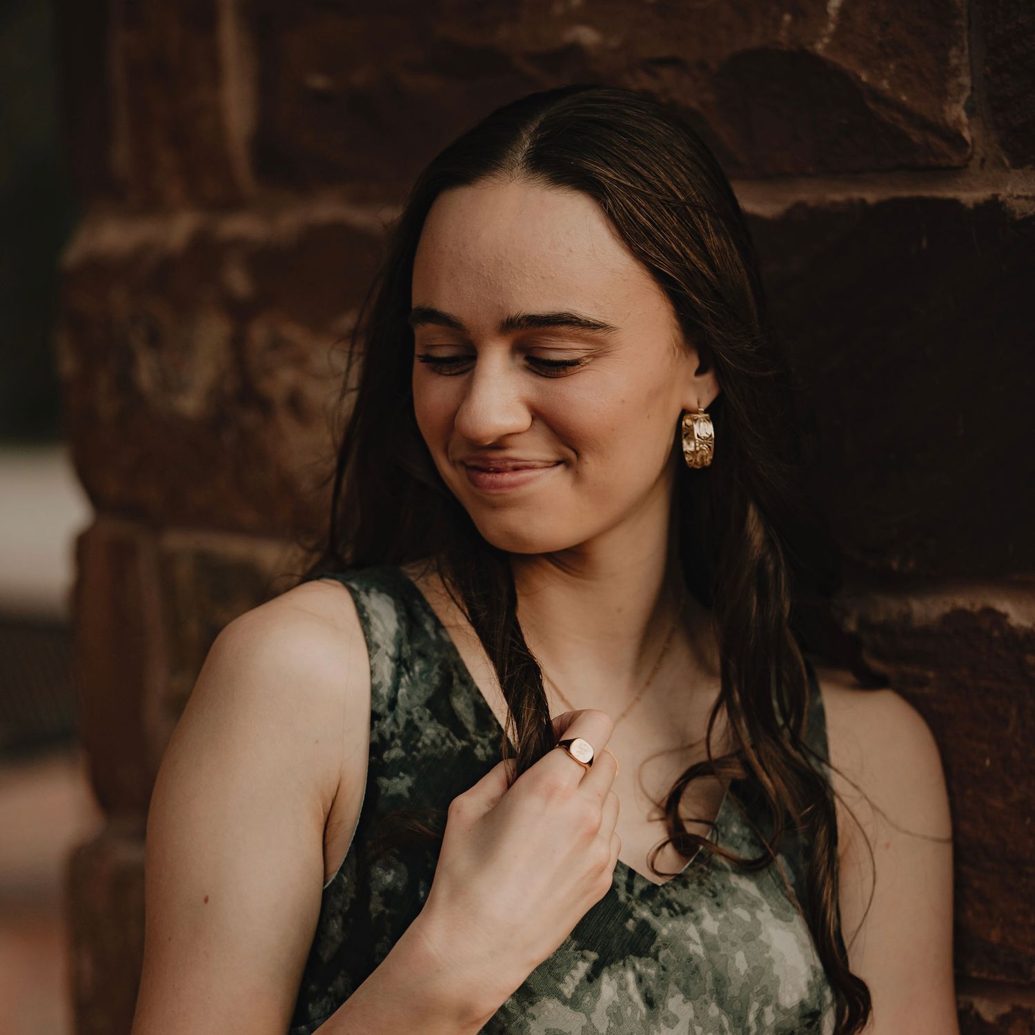 Young woman with long dark hair smiling softly, wearing gold hoop earrings and a green dress, leaning against a brick wall.