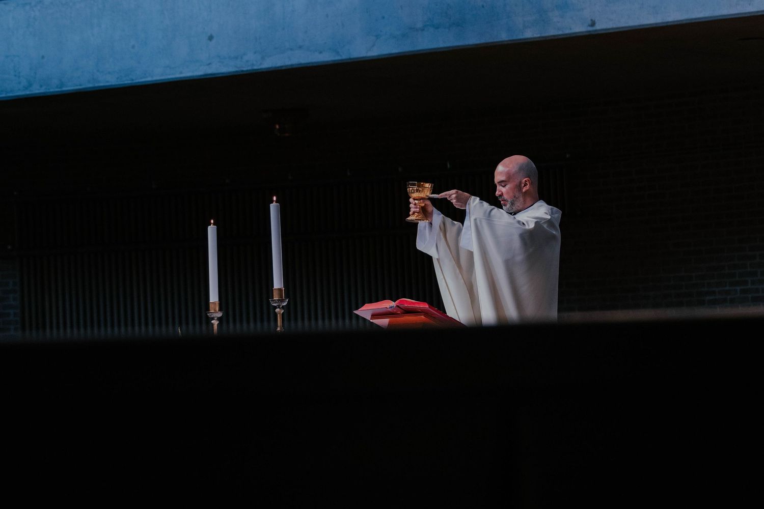A priest in white vestments elevates a chalice during Mass, flanked by candles on an altar.
