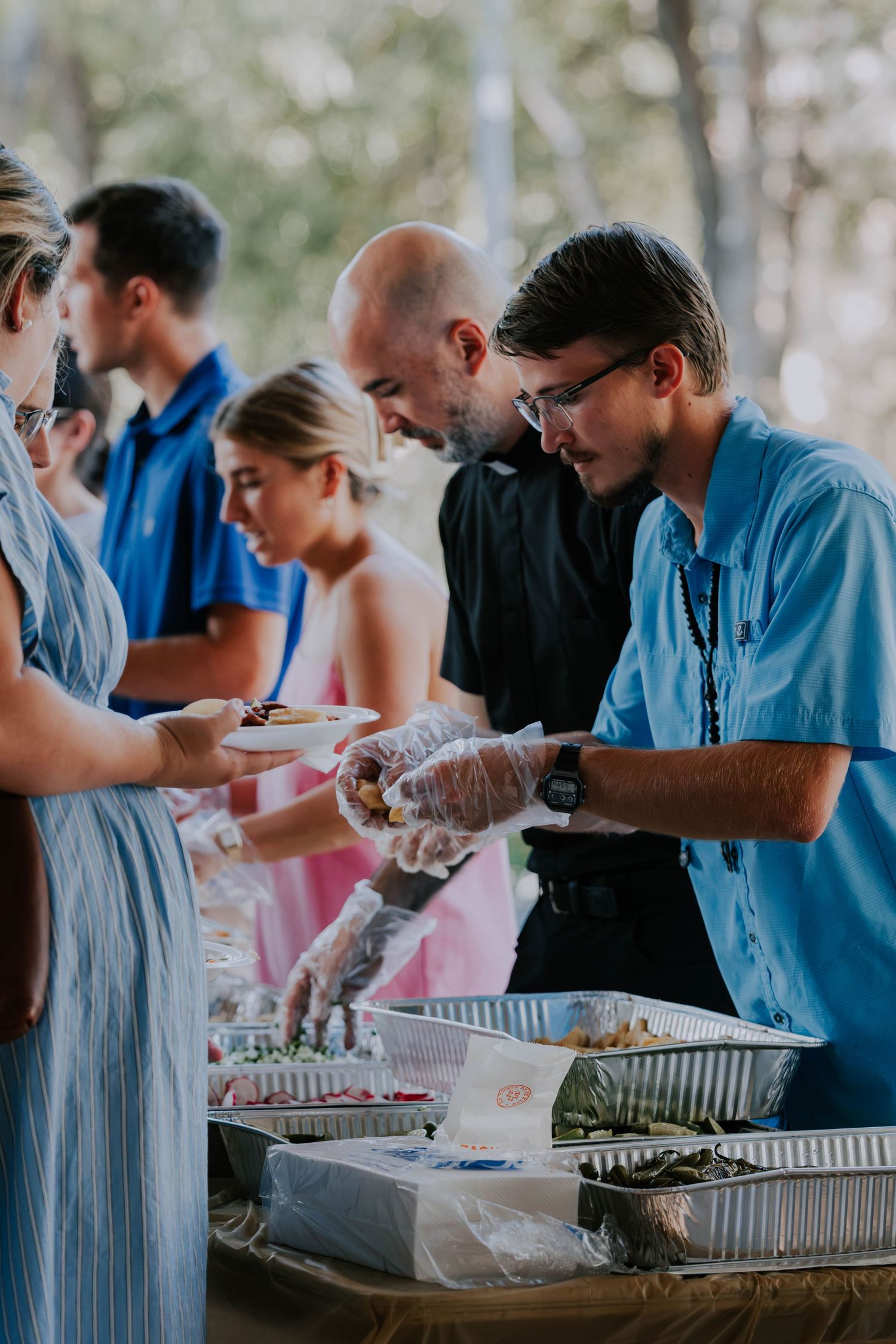 A young man in blue shirt and gloves serves food at an outdoor buffet line during a catered event.