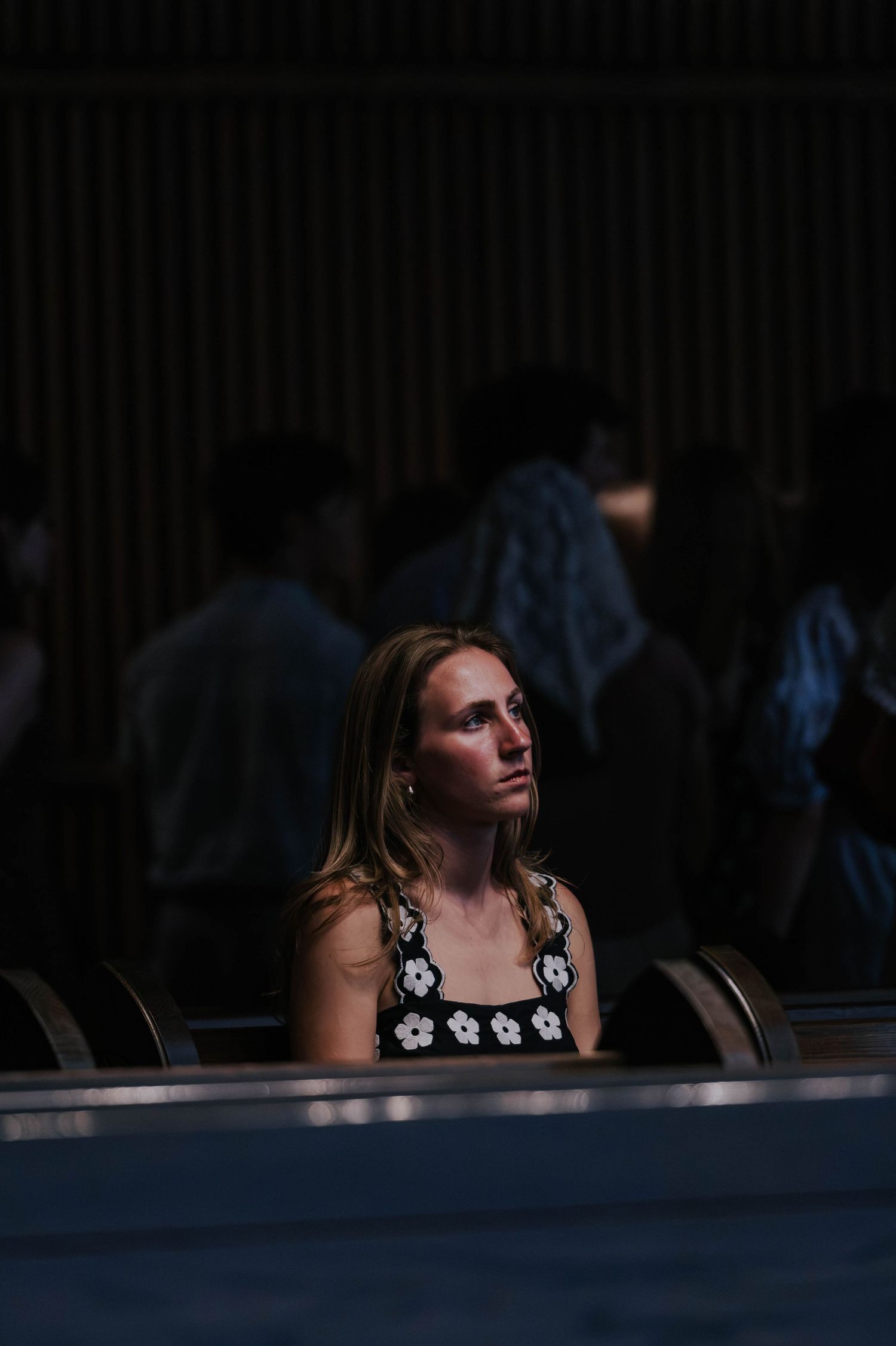 Young woman in black floral crochet top sitting alone in dark venue, looking upward thoughtfully.