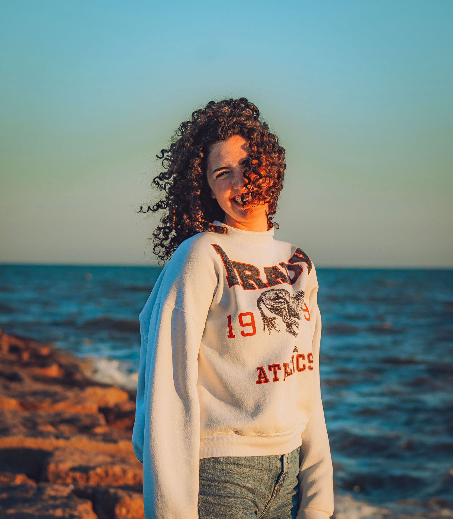 Young woman with curly hair wearing a white athletics sweatshirt posing on a rocky beach at sunset.