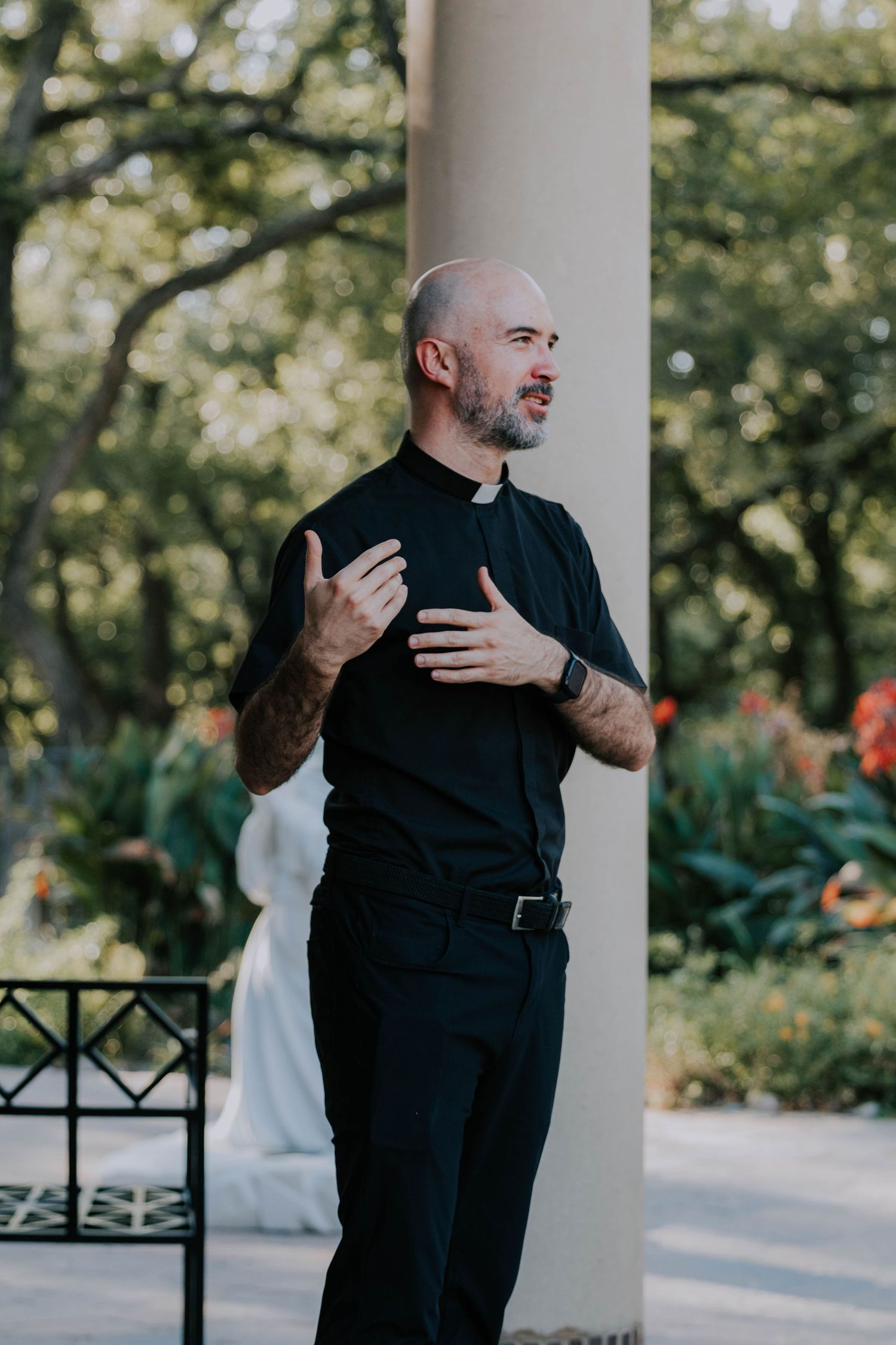 Tattooed Catholic priest in black clerical collar gestures while speaking outdoors near a column in a garden setting.