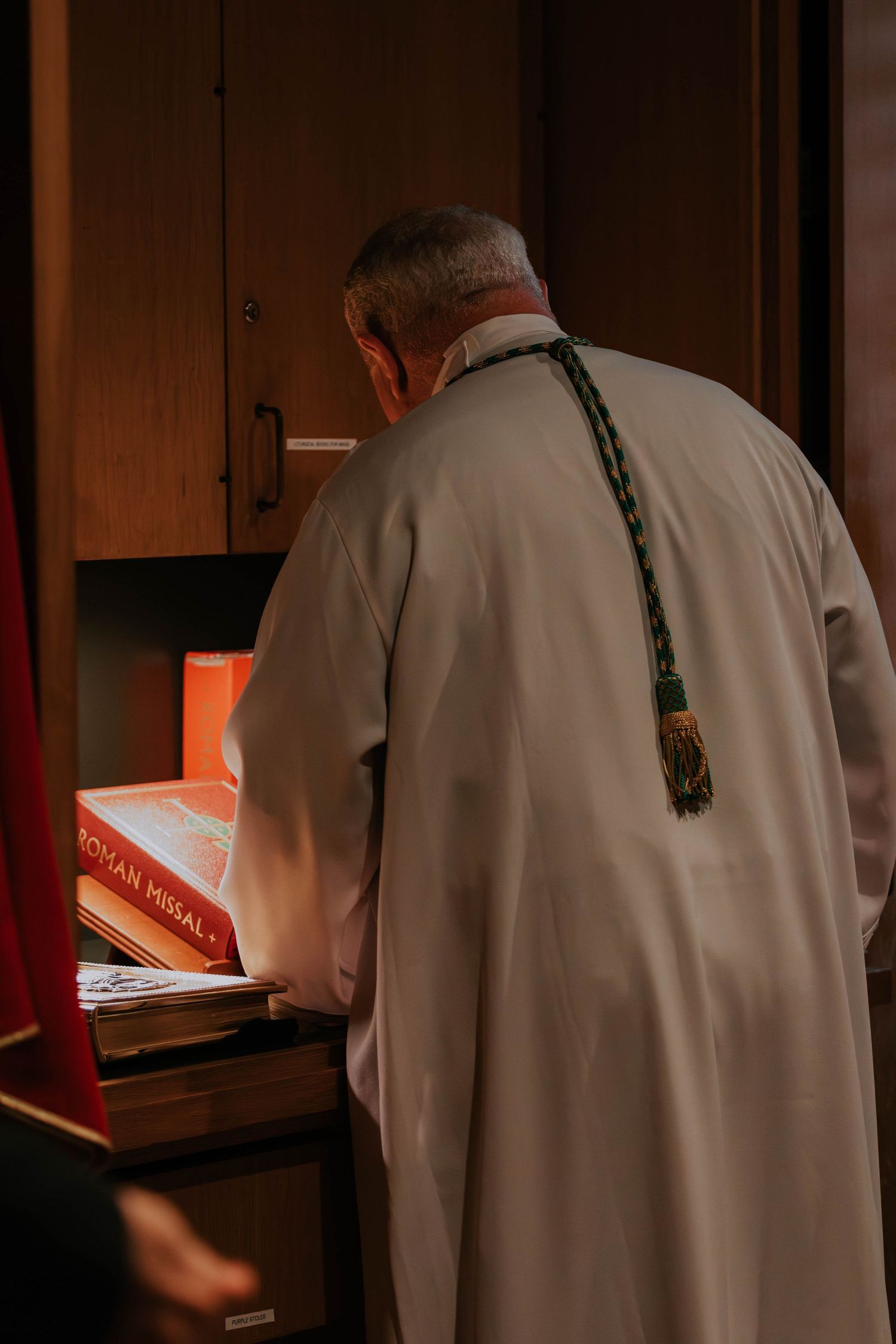Elderly Catholic priest in white vestments with green stole reads Roman Missal at wooden cabinet in dim church sacristy.