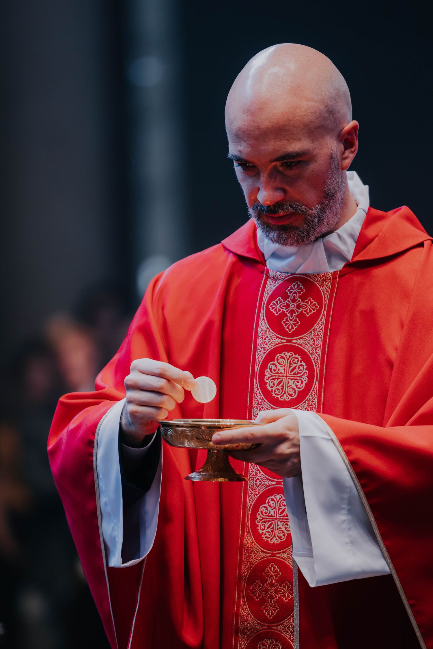 Bald Catholic priest in red vestments holds Eucharist communion wafer during Mass ceremony.