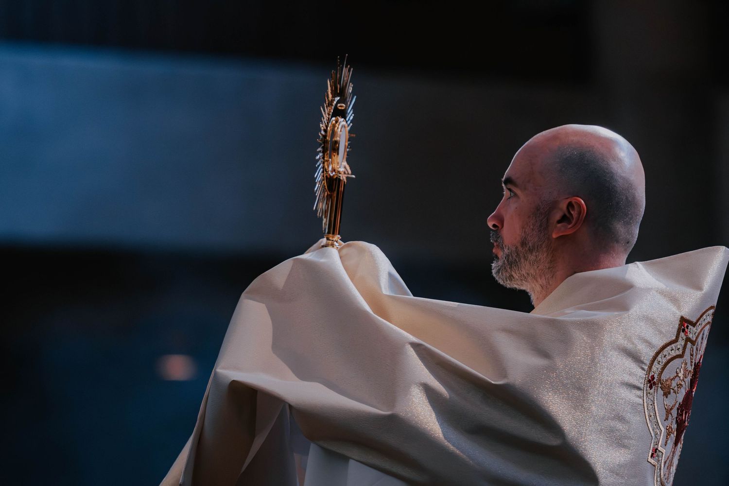 Catholic priest in white vestments raises a monstrance during a religious ceremony.
