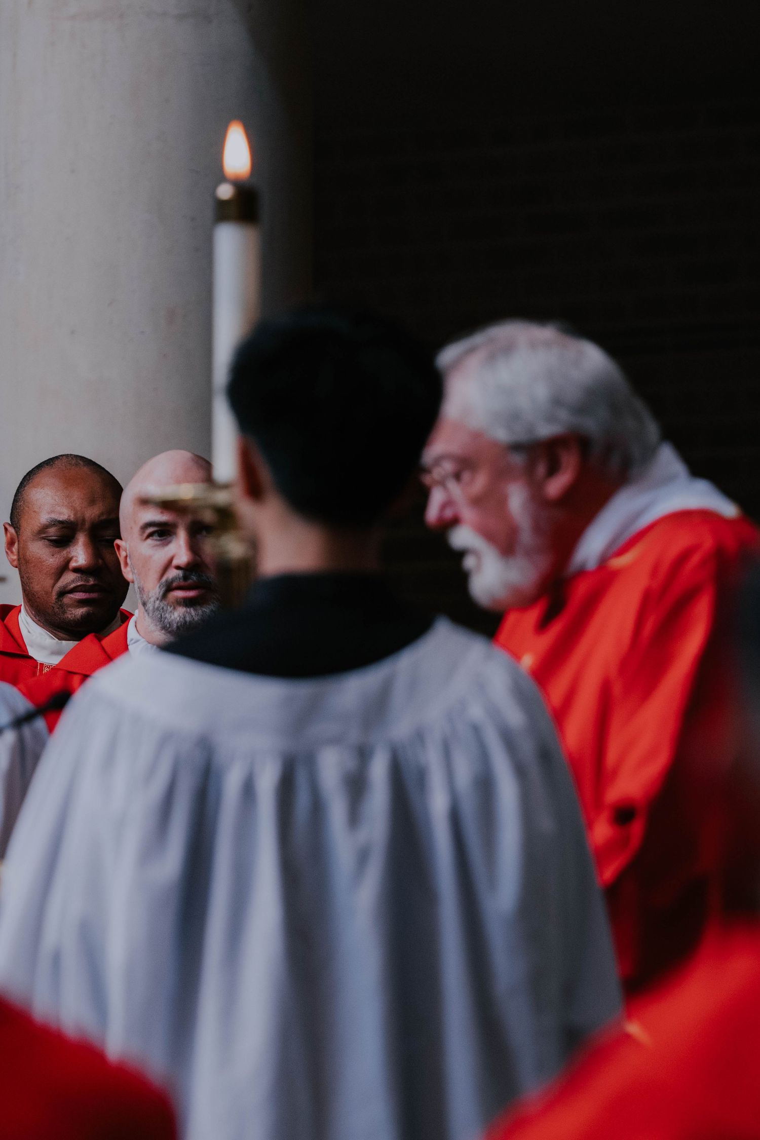 Clergy members in red and white robes gather around a lit candle during a religious ceremony.