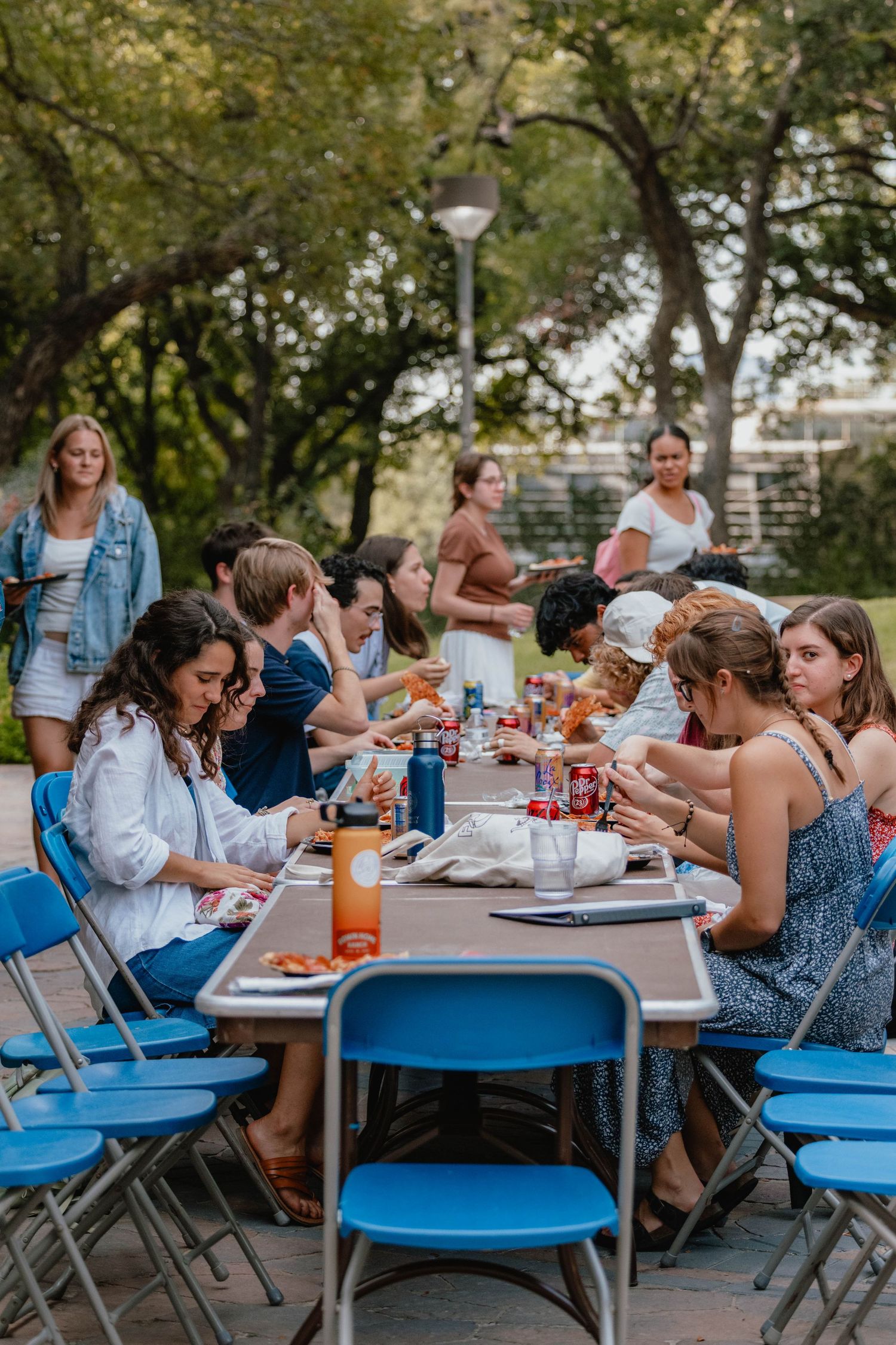 Group of young adults enjoying an outdoor meal together at long tables with blue chairs in a park setting.