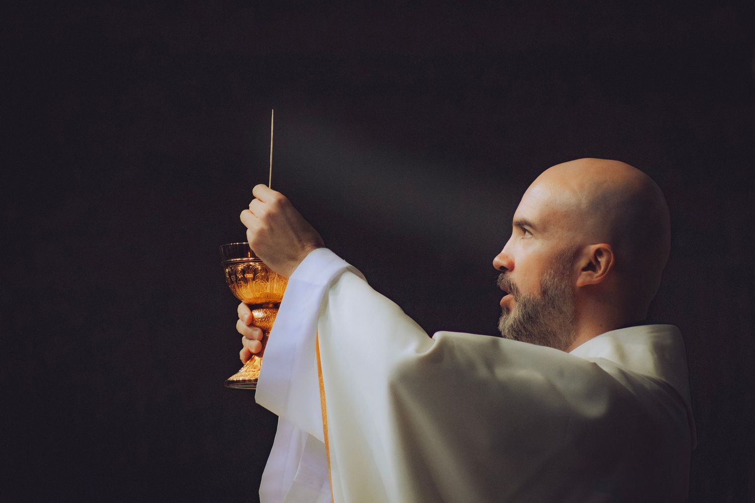 Bald Catholic priest in white vestments elevating a golden chalice during Mass in dramatic lighting.