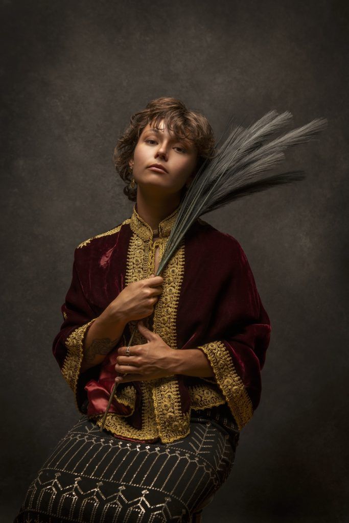 Young person in ornate Renaissance-style costume holding dried wheat stalks in dramatic portrait.