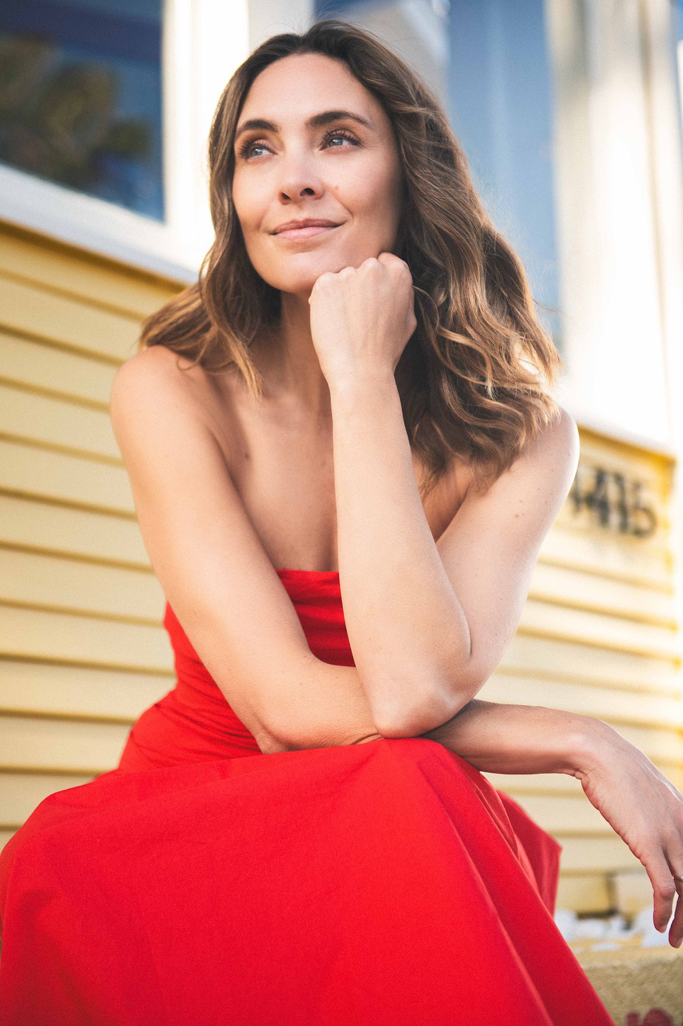 A woman in a red strapless dress sits gracefully outdoors, resting her chin on her hand with a gentle smile.