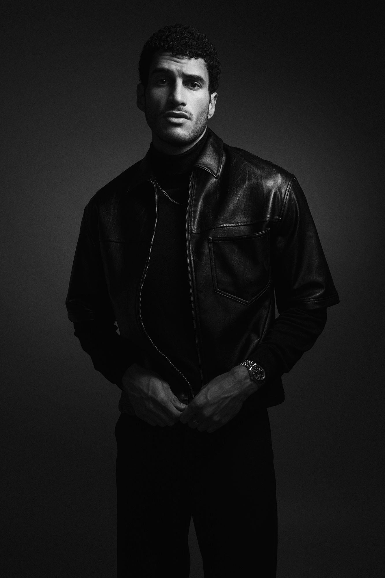 Black and white portrait of a male model in a leather jacket and turtleneck on dark background.