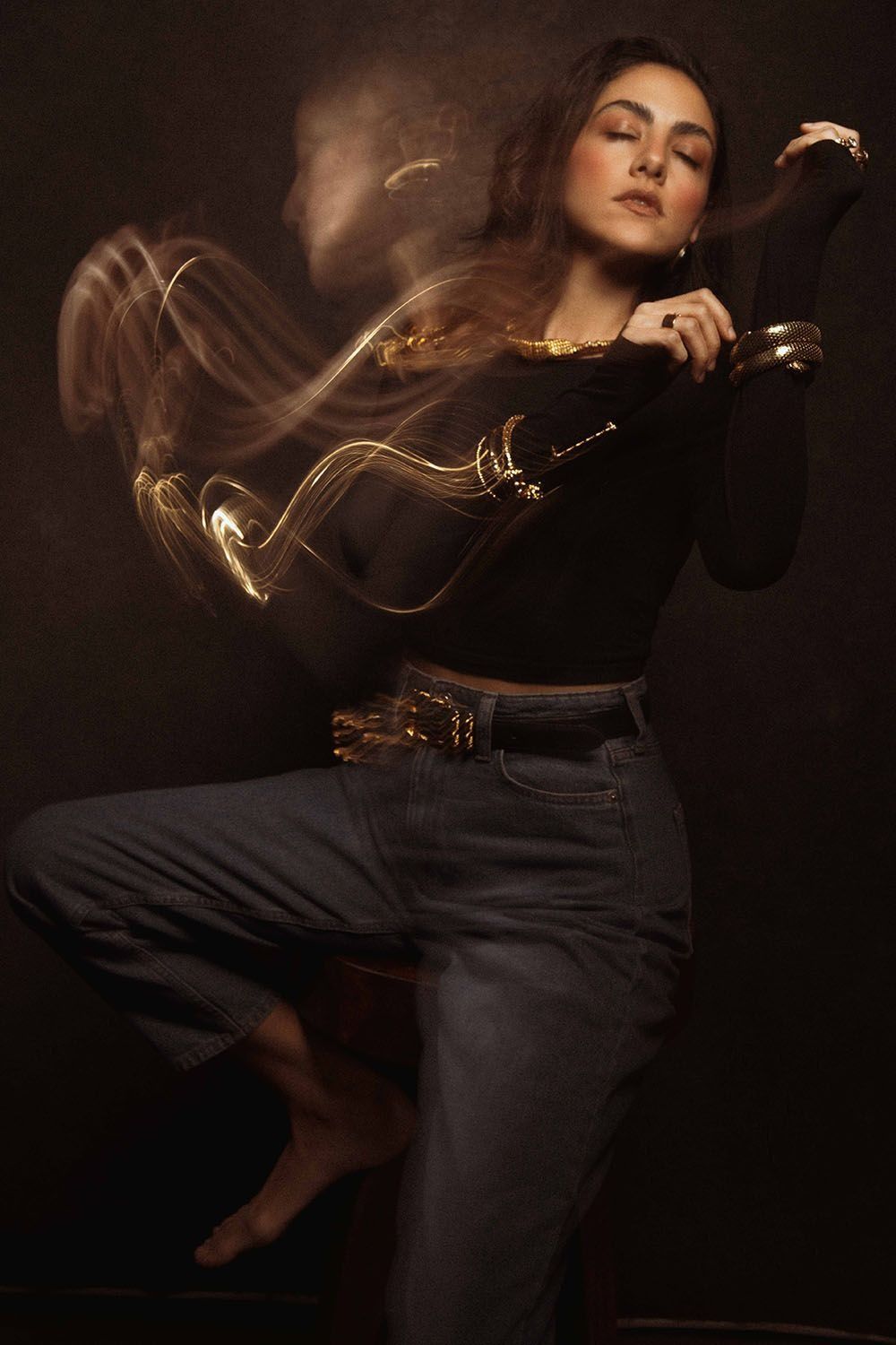 A woman in black crop top and jeans poses dynamically with golden light trails swirling around her in a dark studio.