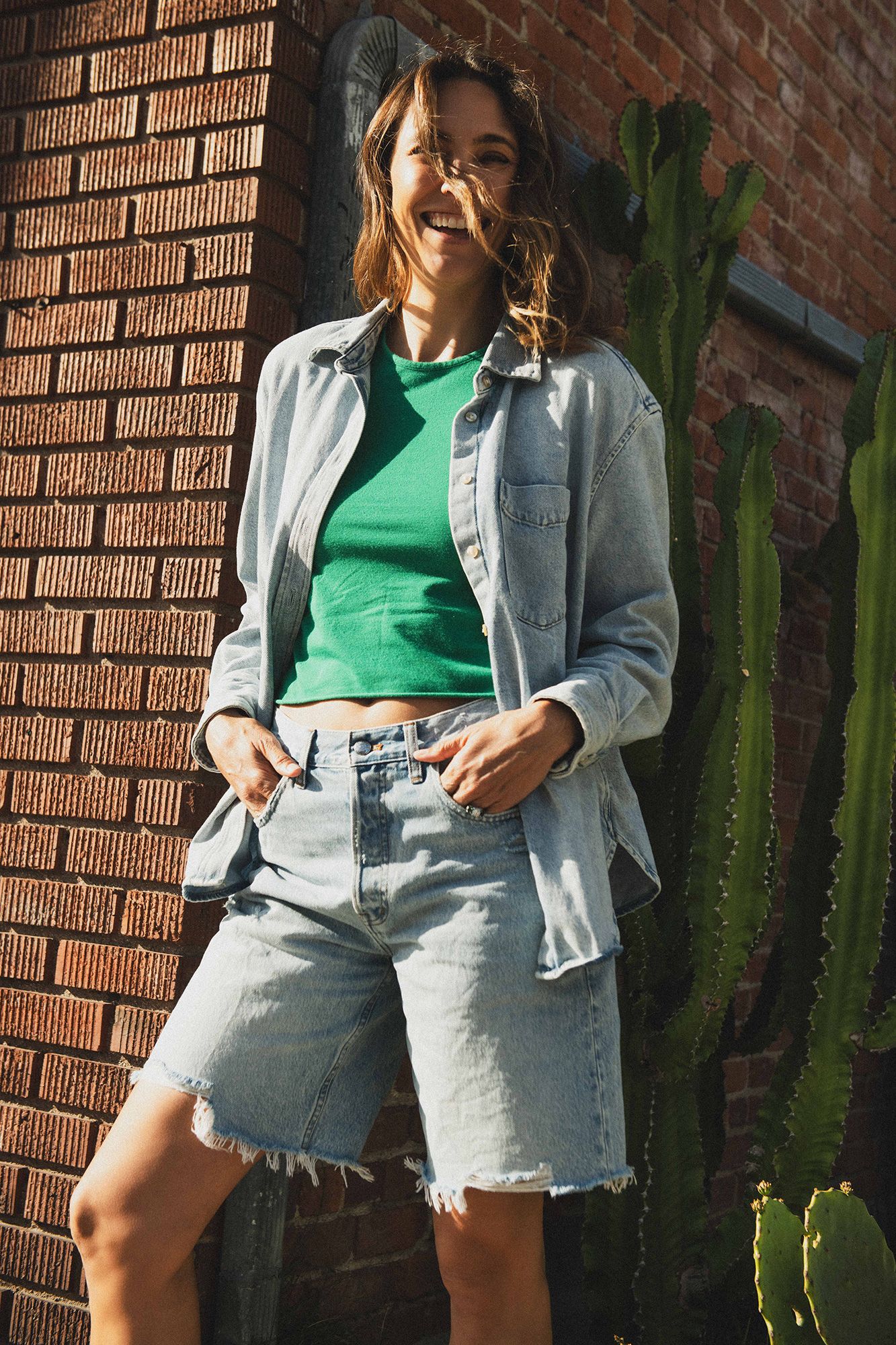 Woman in denim jacket, green crop top, and cutoff jean shorts poses against a brick wall with cacti.