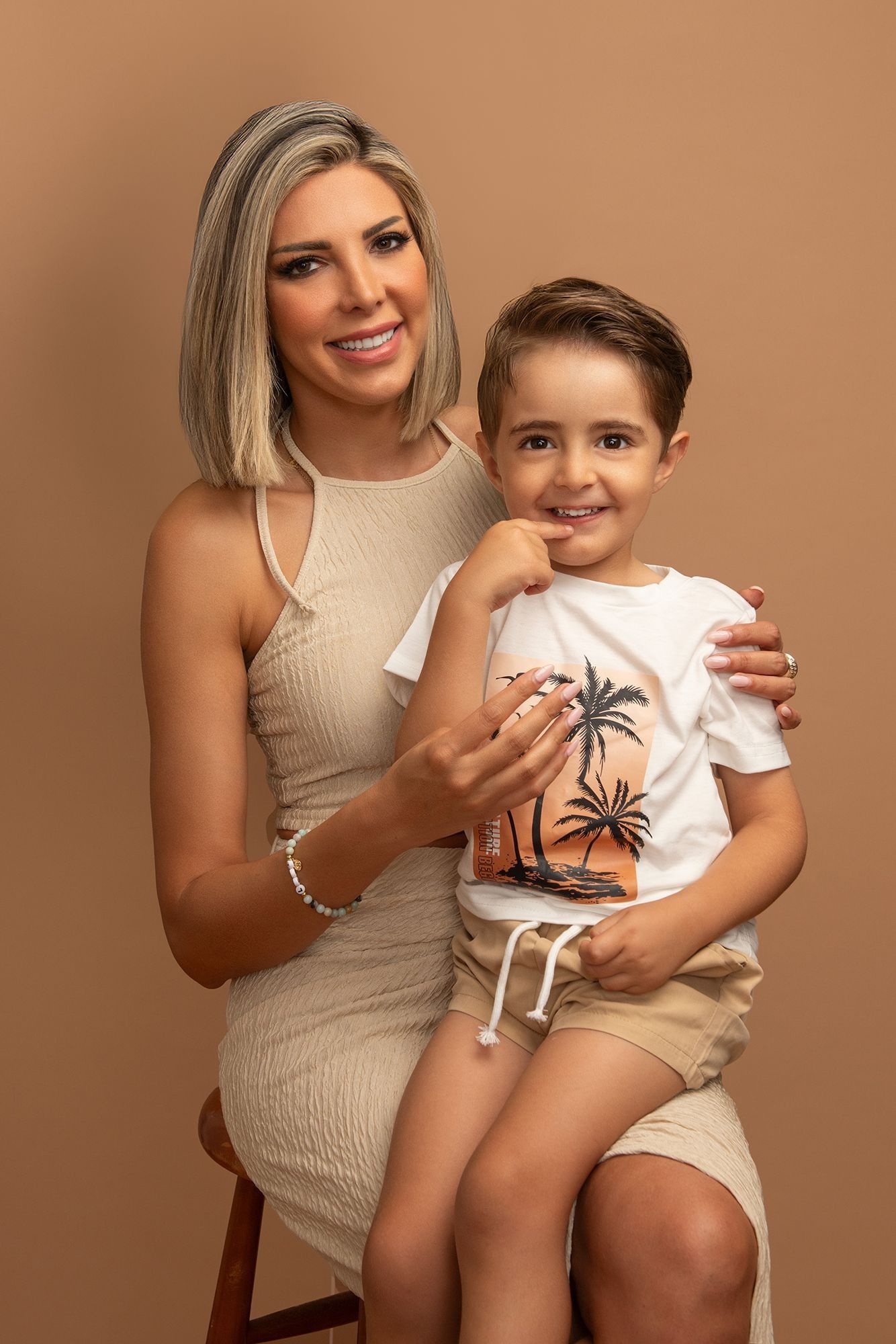 Smiling woman in beige dress sitting with young boy on her lap against tan background.