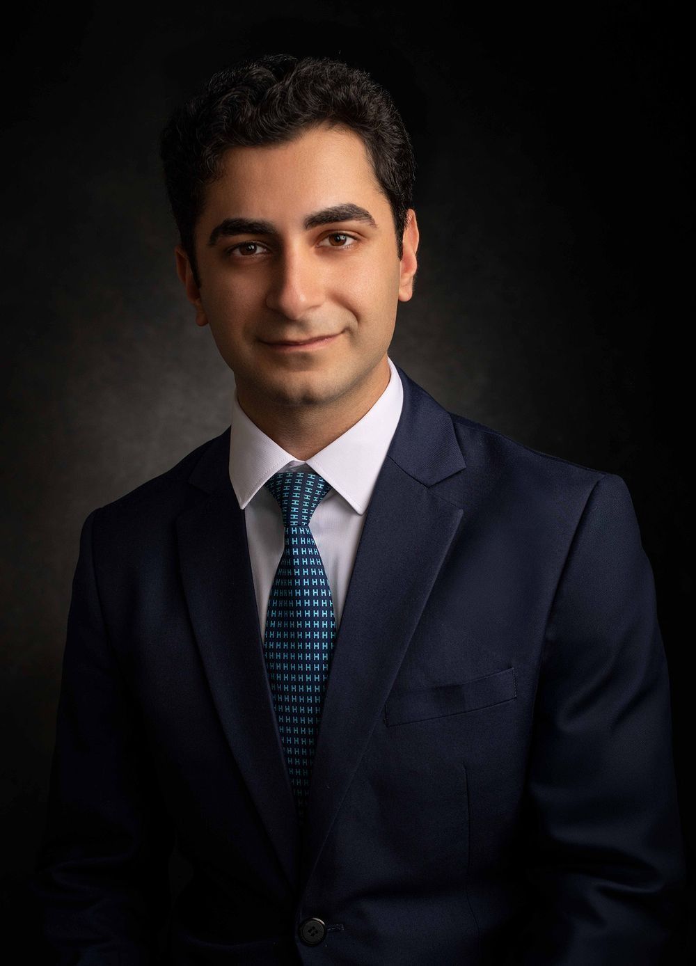 Professional headshot of a young man in a navy suit and teal tie against a dark background.