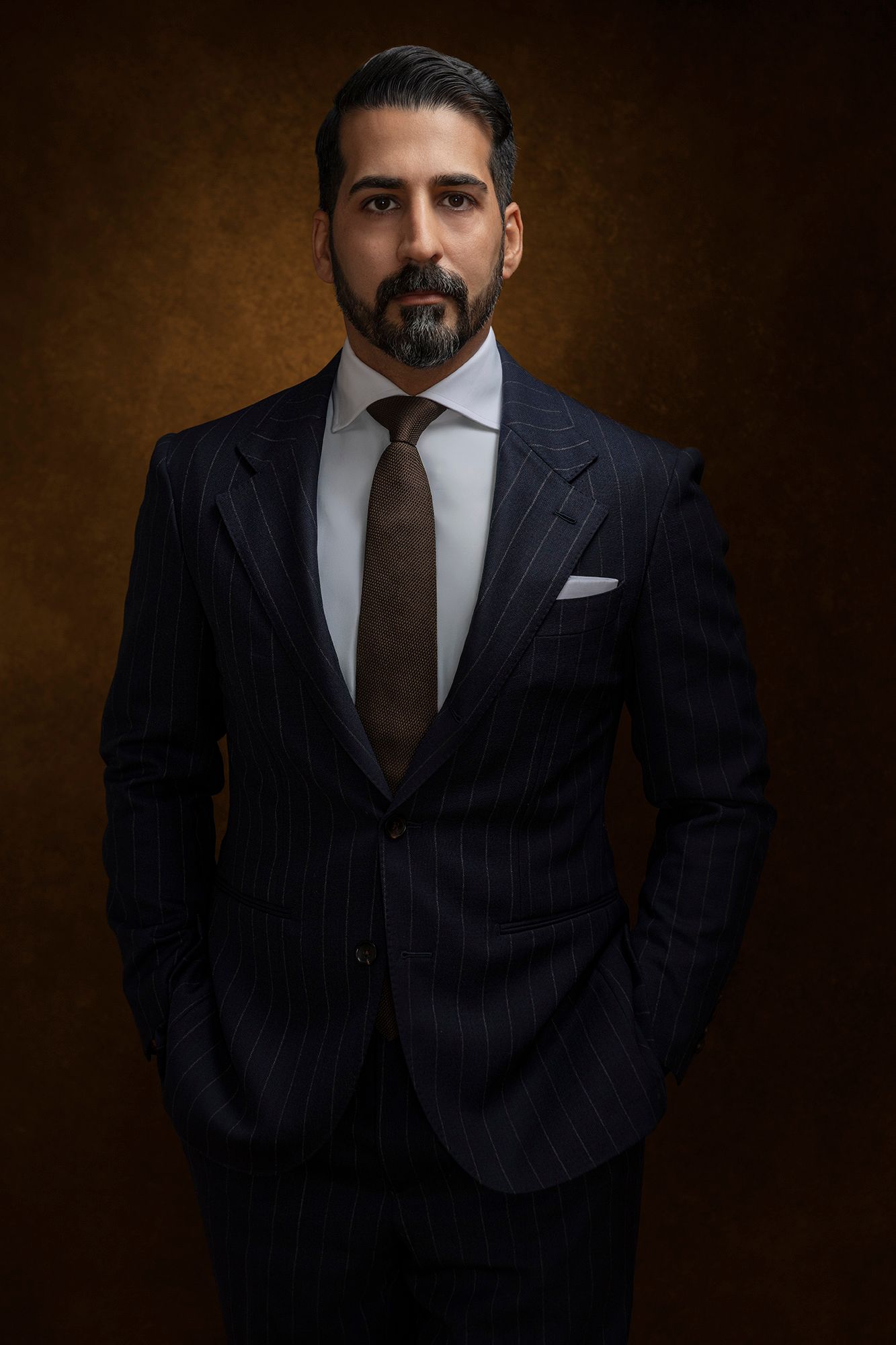 Bearded man in navy pinstripe suit with tie and pocket square posing against brown background.