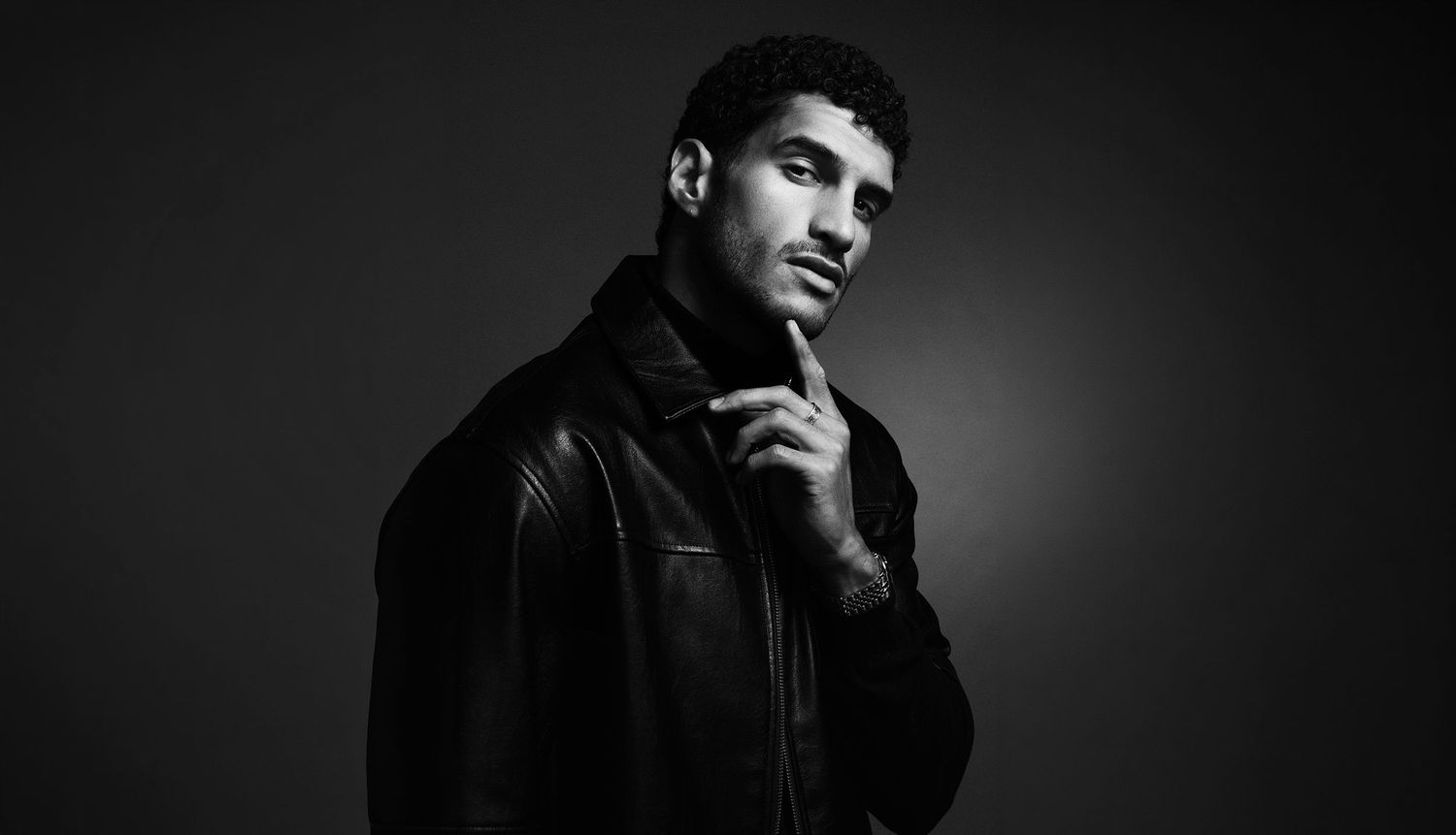 Man in black leather jacket posing thoughtfully with hand on chin in black and white studio portrait.