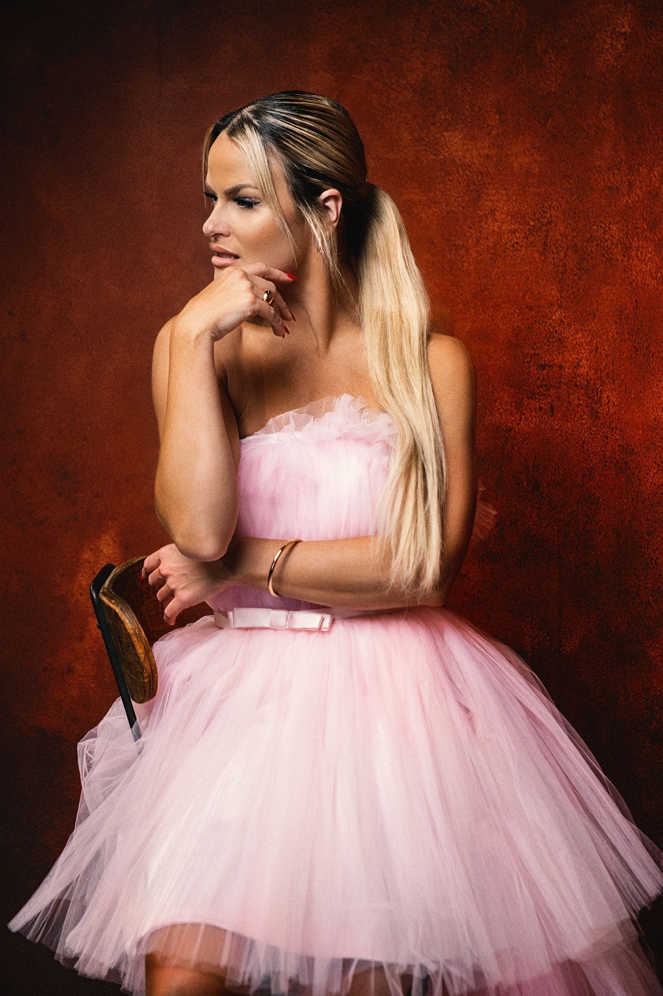 Blonde woman in pink tulle dress holding shoe against red background.