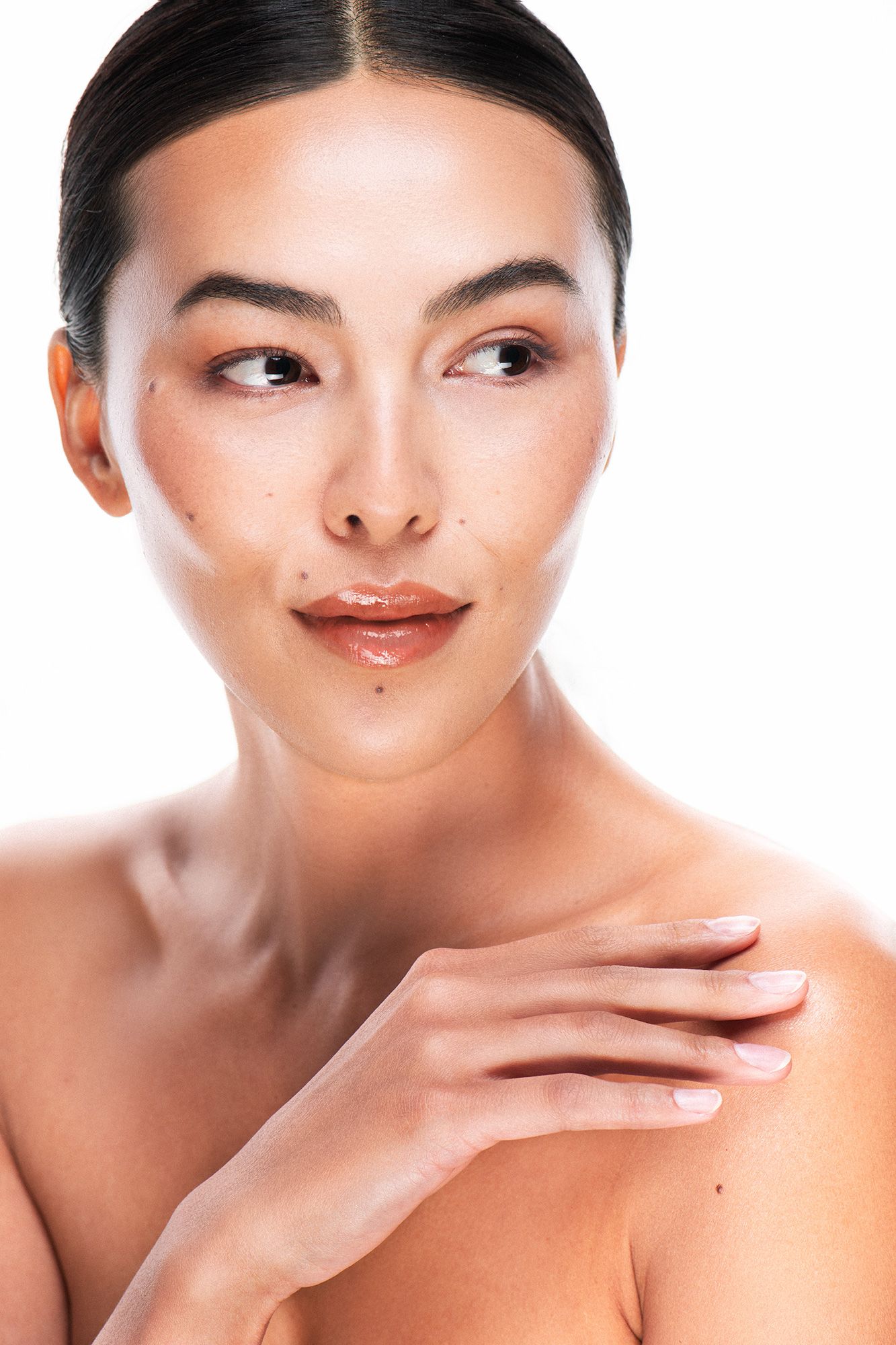 Young Asian woman with natural makeup touches her shoulder, gazing sideways against white background.