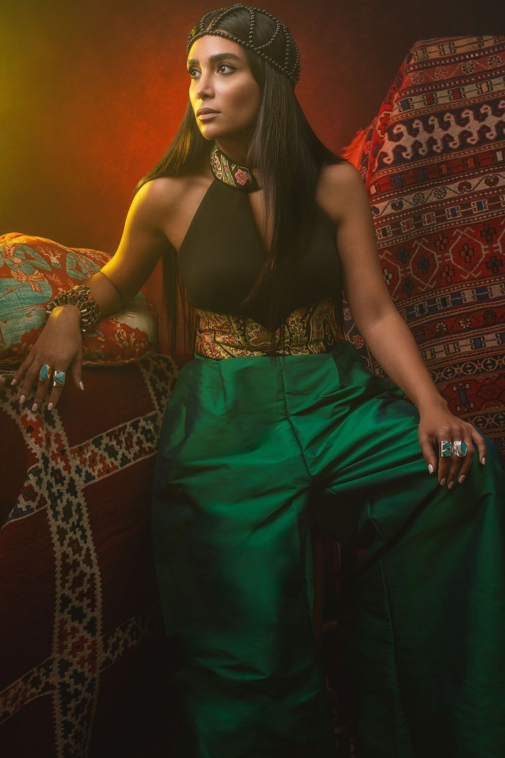 Woman in Egyptian-inspired costume with green pants and gold accessories seated on colorful cushions.